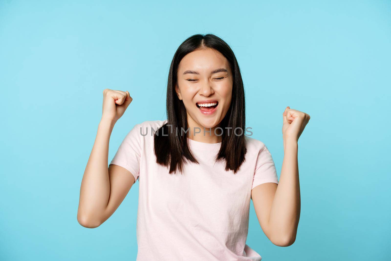 Happy satisfied korean girl winning, celebrating victory, triumphing with fist pump, scream of joy, standing over blue background.