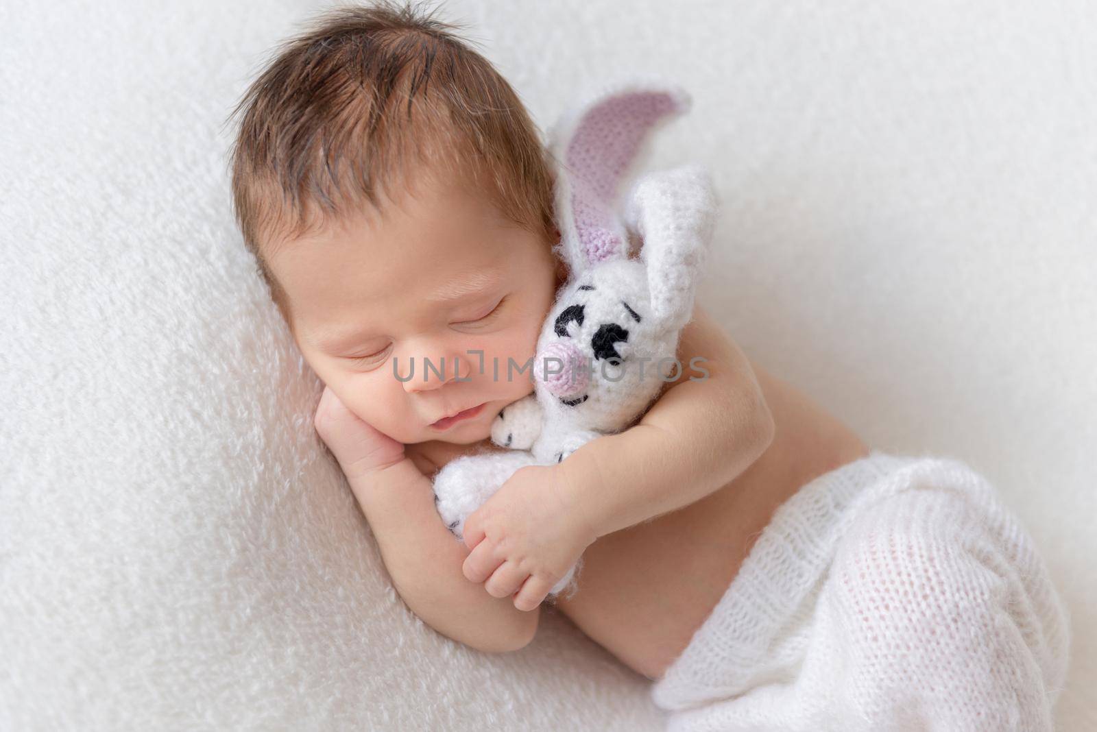 Little cute baby in white knitted pants holding white toy bunny and sleeping on white soft bedcover