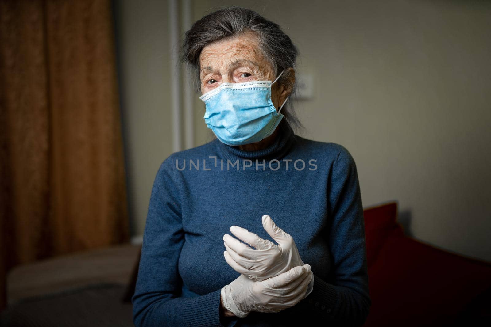 Senior woman in protective medical mask and latex gloves stands and watches into camera with sad, tired, upset emotions at home. Theme lockdown, stay home, coronavirus and covid 19 by Tomashevska