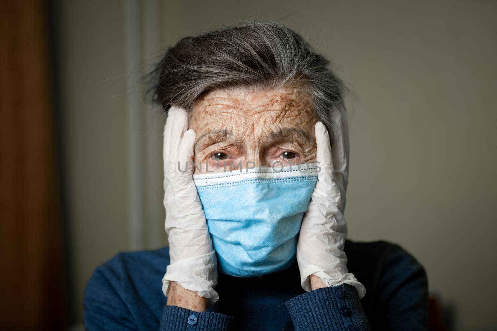 Portrait senior woman holding head hands, sad worried emotion in protective medical mask, gloves against virus and infection at home. Old gray hair female quarantine stay at home, loneliness and fear by Tomashevska