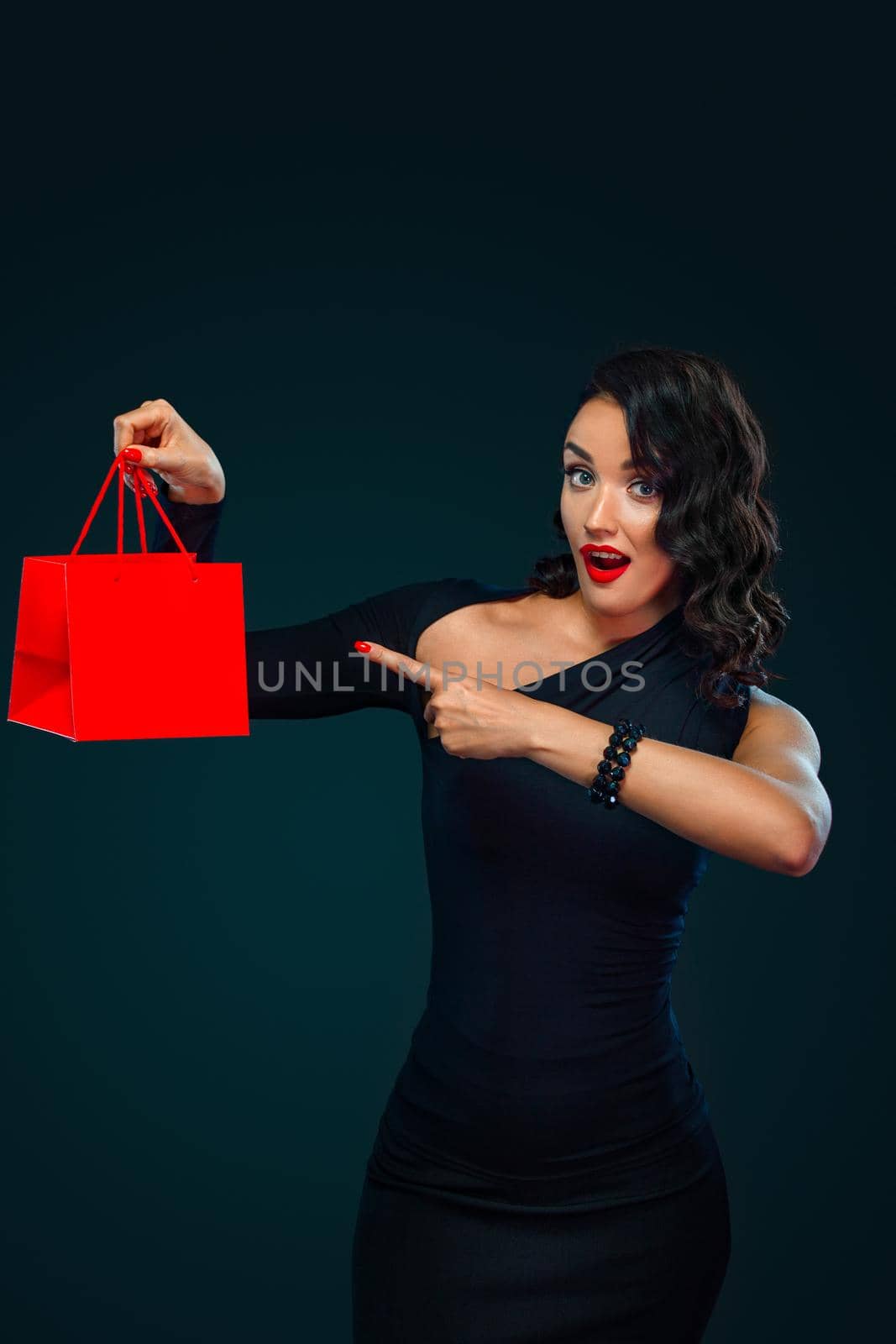 Beautiful young woman make shopping in black friday holiday. Girl with black bag on dark background. Beautiful girl with curly hair pointing to looking left.