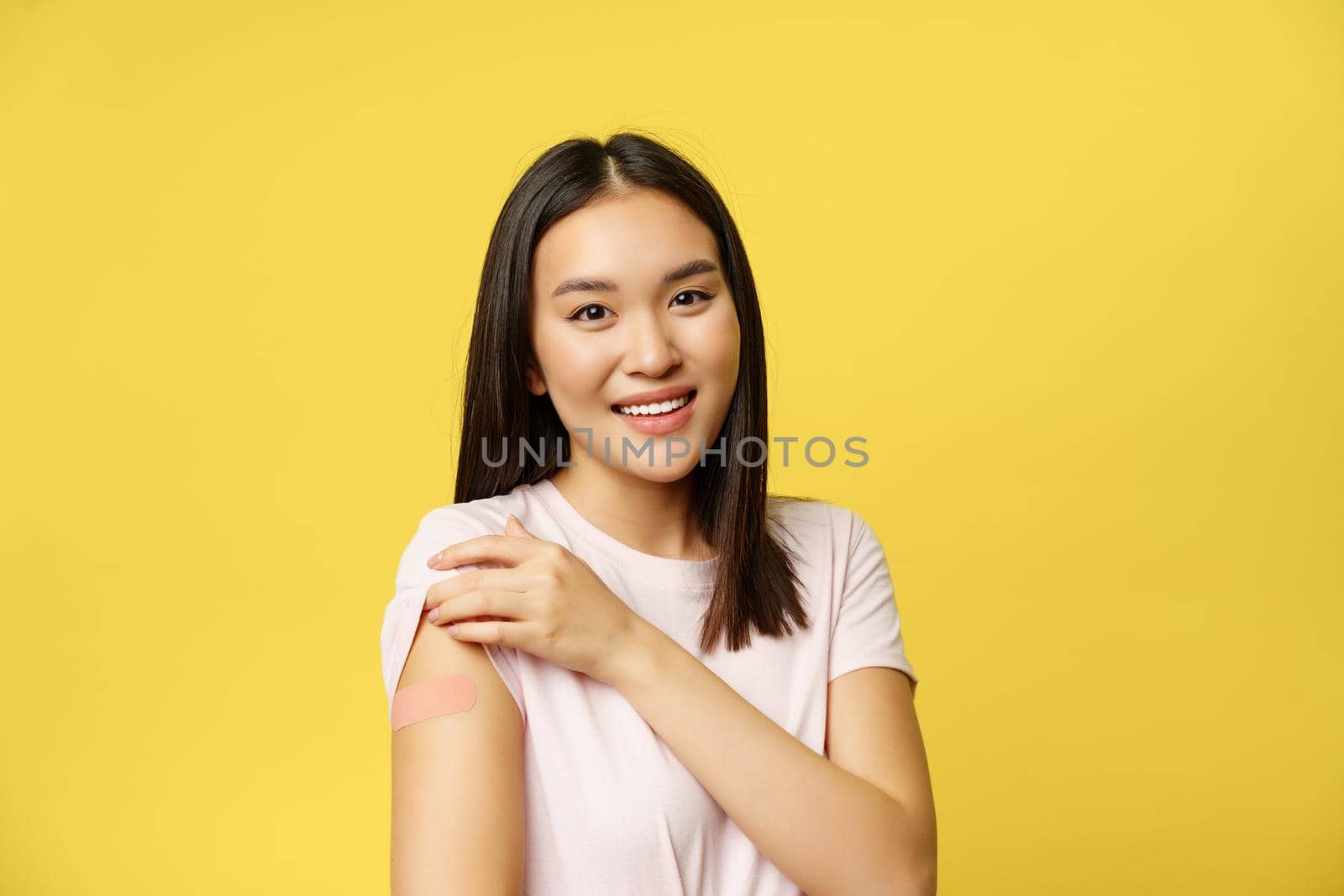 Covid-19 and healthcare medical concept. Smiling woman shows shoulder with patch, vaccinated from coronavirus, standing in t-shirt over yellow background.