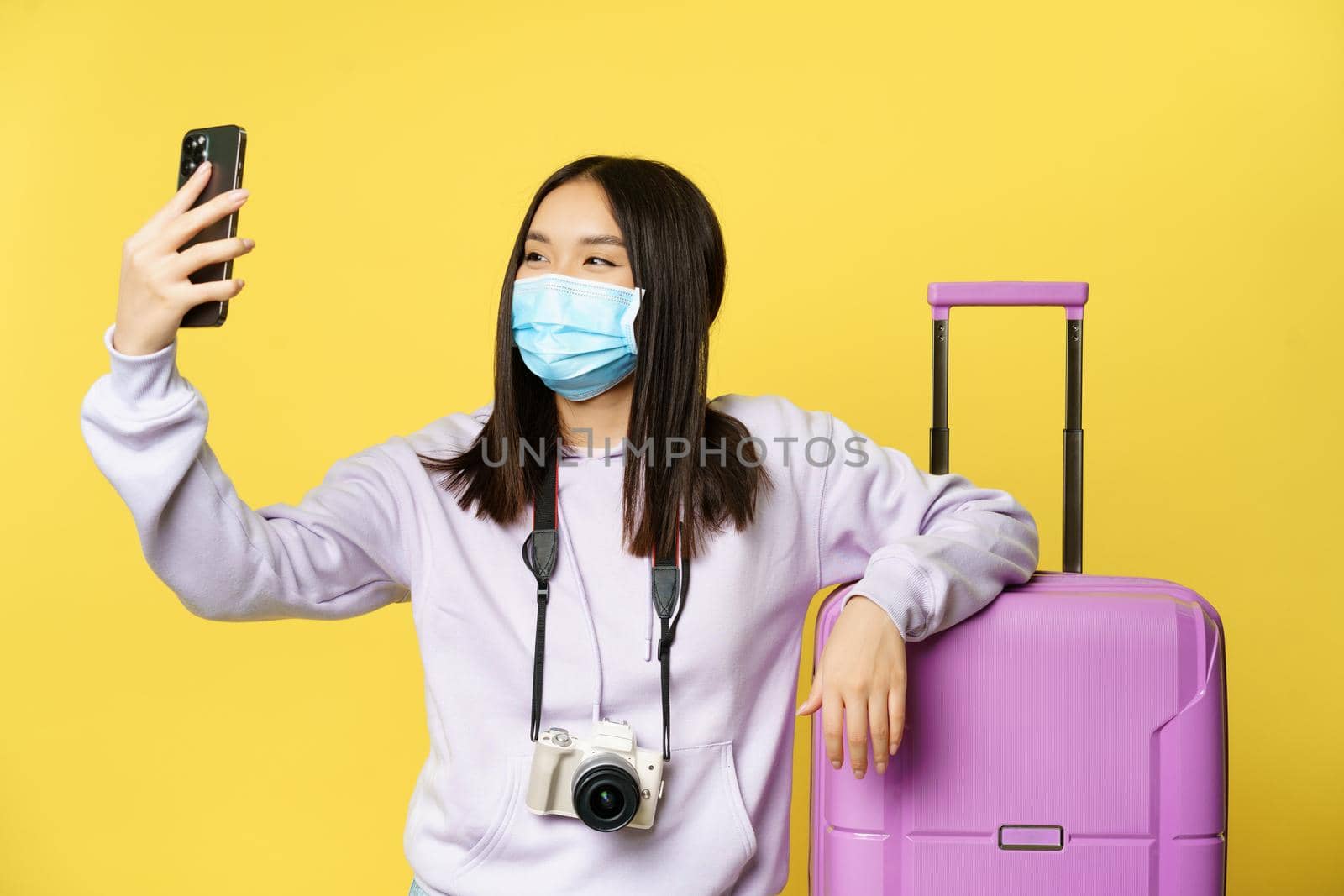 Happy tourist, asian girl on vacation taking selfie in medical mask near suitcase, photographing on smartphone, yellow background.