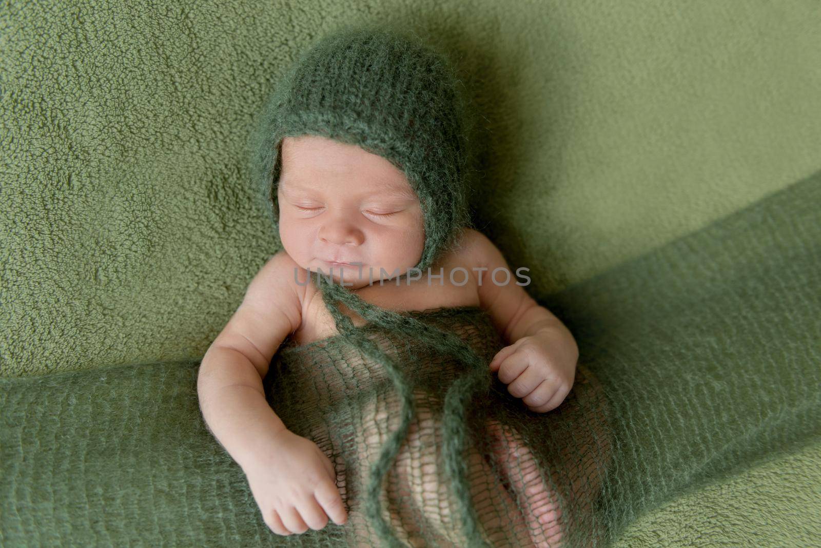 Little baby in dark green knitted beanie covered in lilac blanket sweetly sleeping on the soft green bedcover