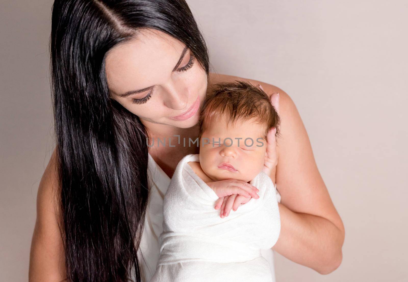Mother holding little sleeping baby covered in white coverlet on the light background