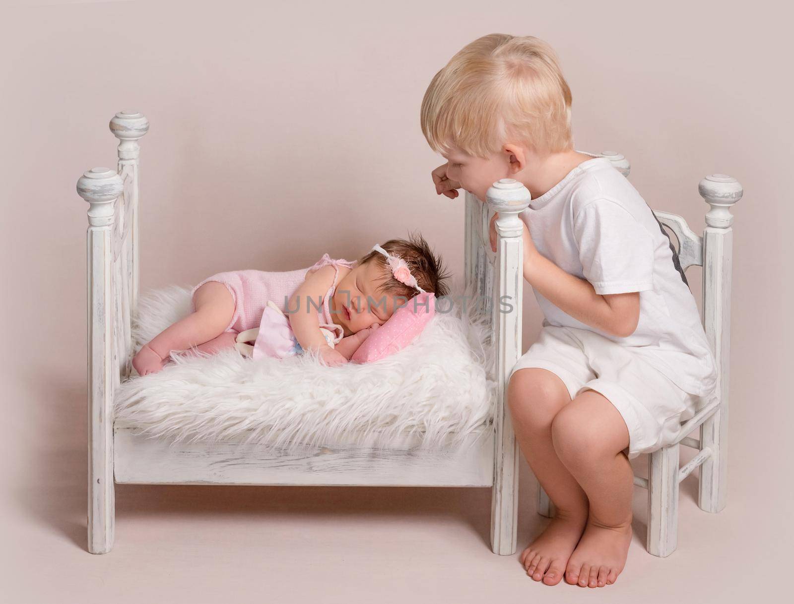 Little blond boy looking on baby in pink knitted suit sweetly sleeping in tiny bed with soft white blanket
