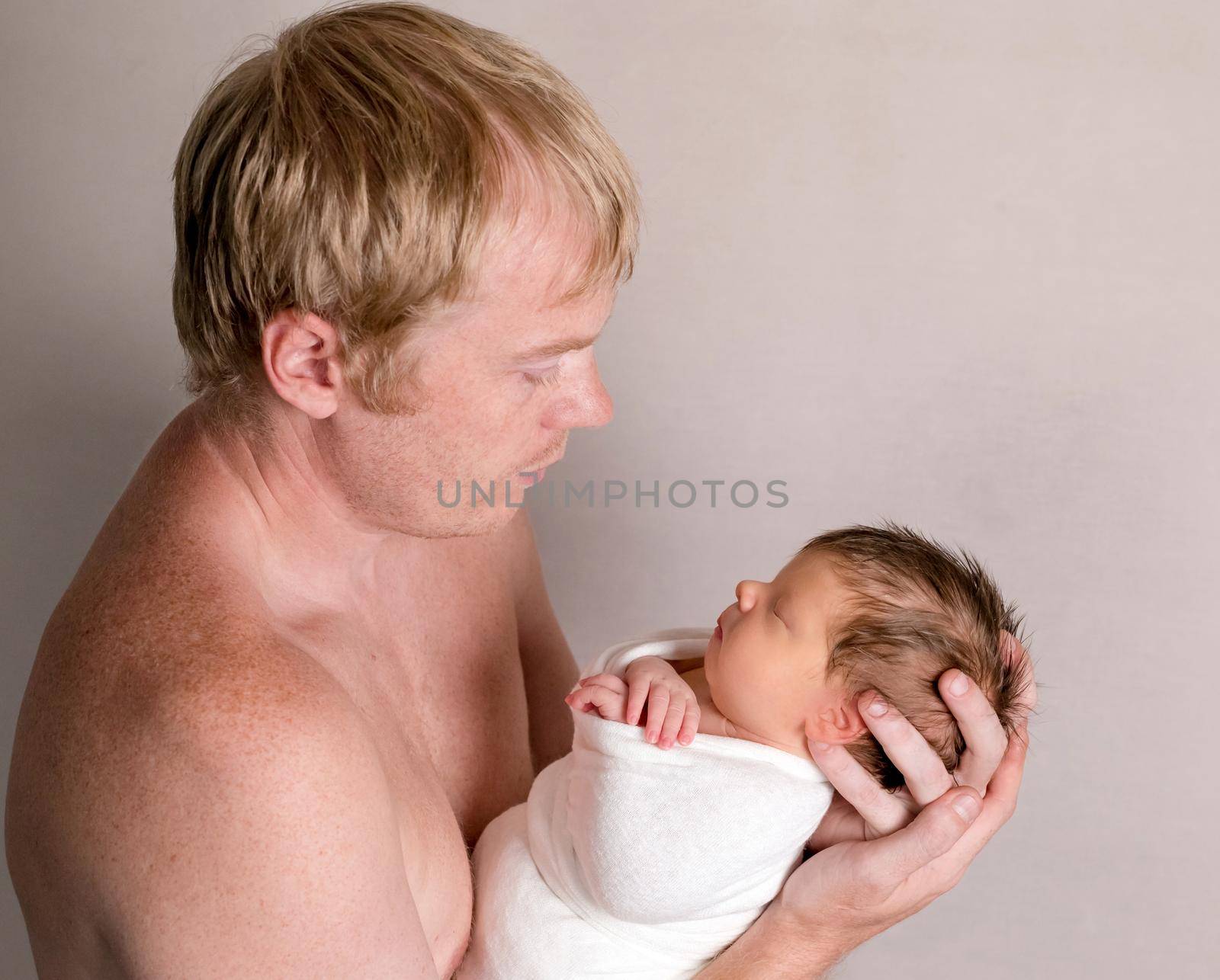 Father holding and looking on little sleeping baby covered in white coverlet on the light background