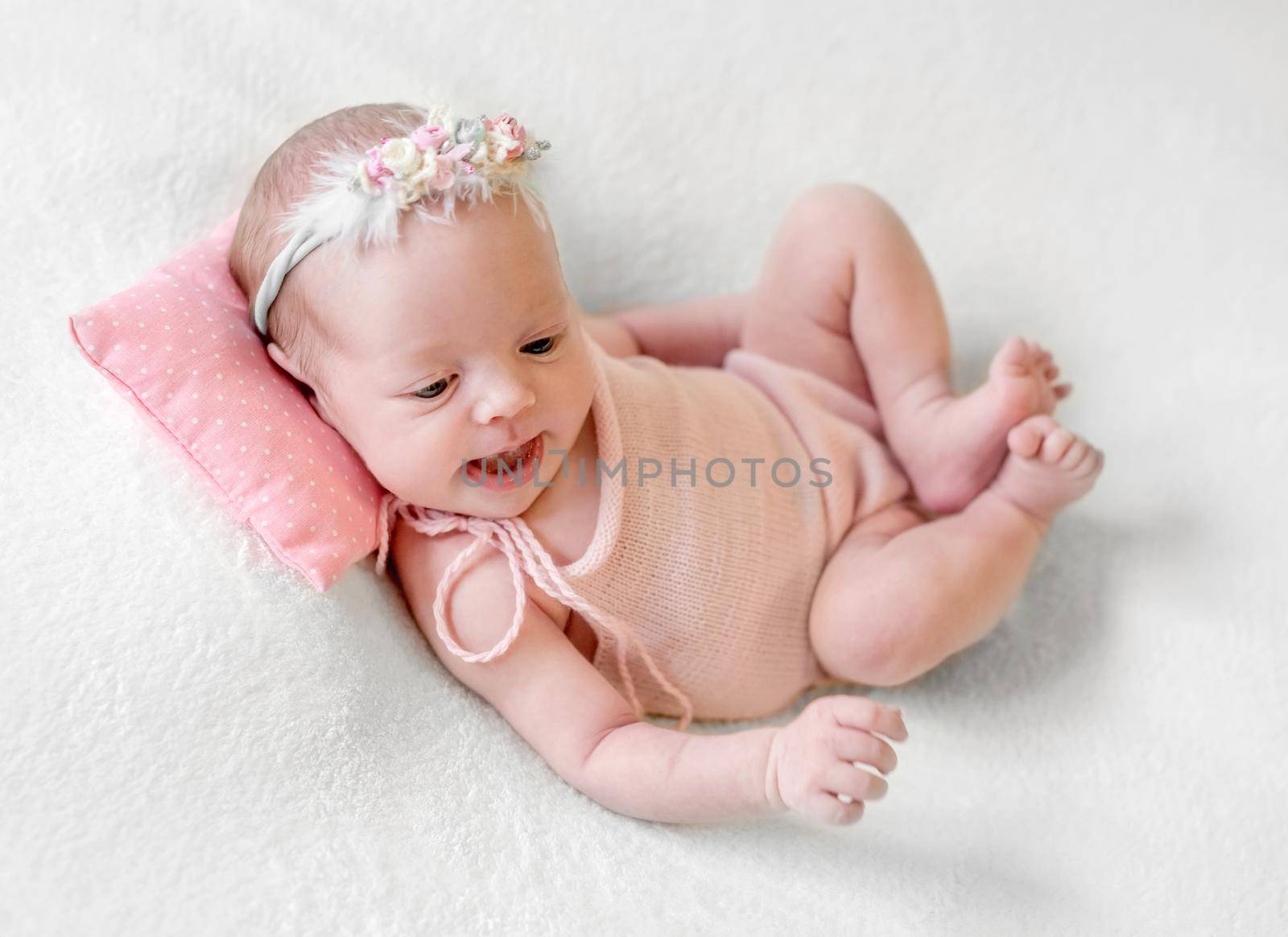 Little smiling baby in pink knitted suit joyfully lying on pink dotted pillow and white soft blanket