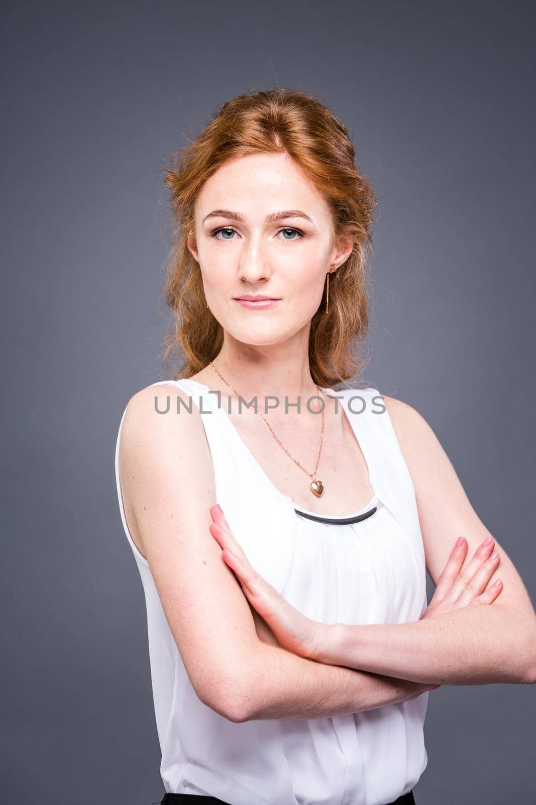 portrait of a young redhaired beautiful girl in the studio on a gray isolated background. A woman is standing with her arms folded and smiling in a white shirt with a short sleeve. Business concept by Tomashevska