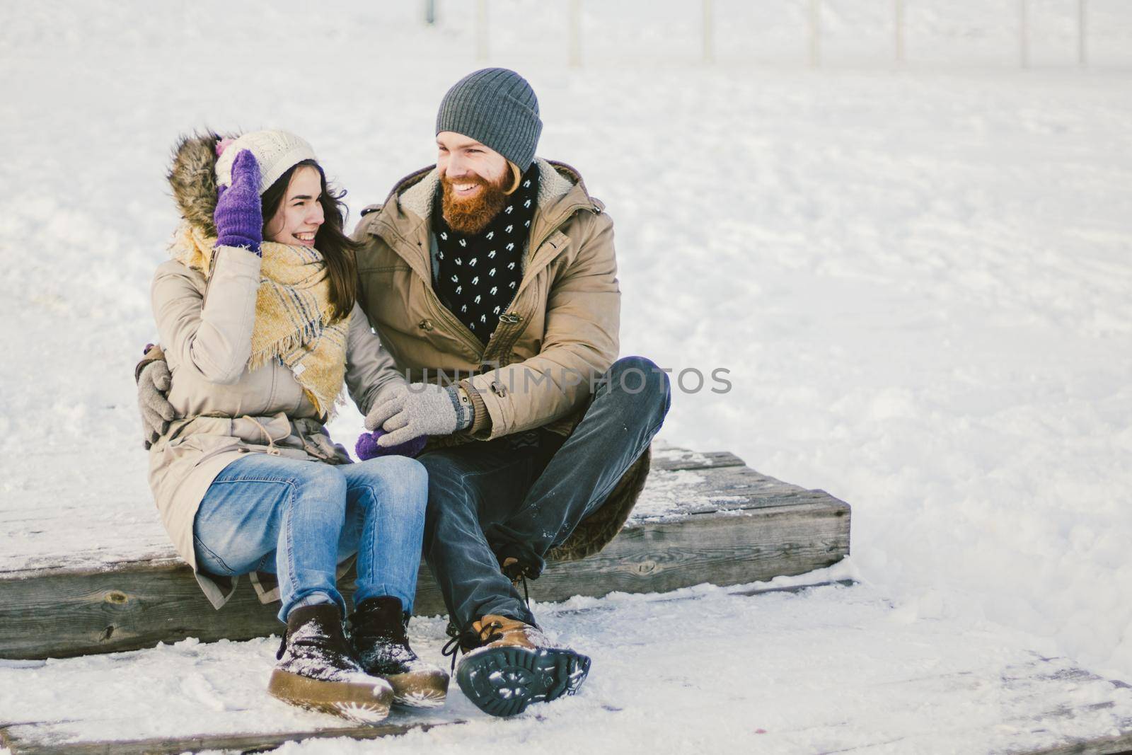 Loving couple young people a man with a beard and a Caucasian woman are sitting on a wooden staircase in the winter on the snow St. Valentine's Day a date Christmas hugging and happy by Tomashevska