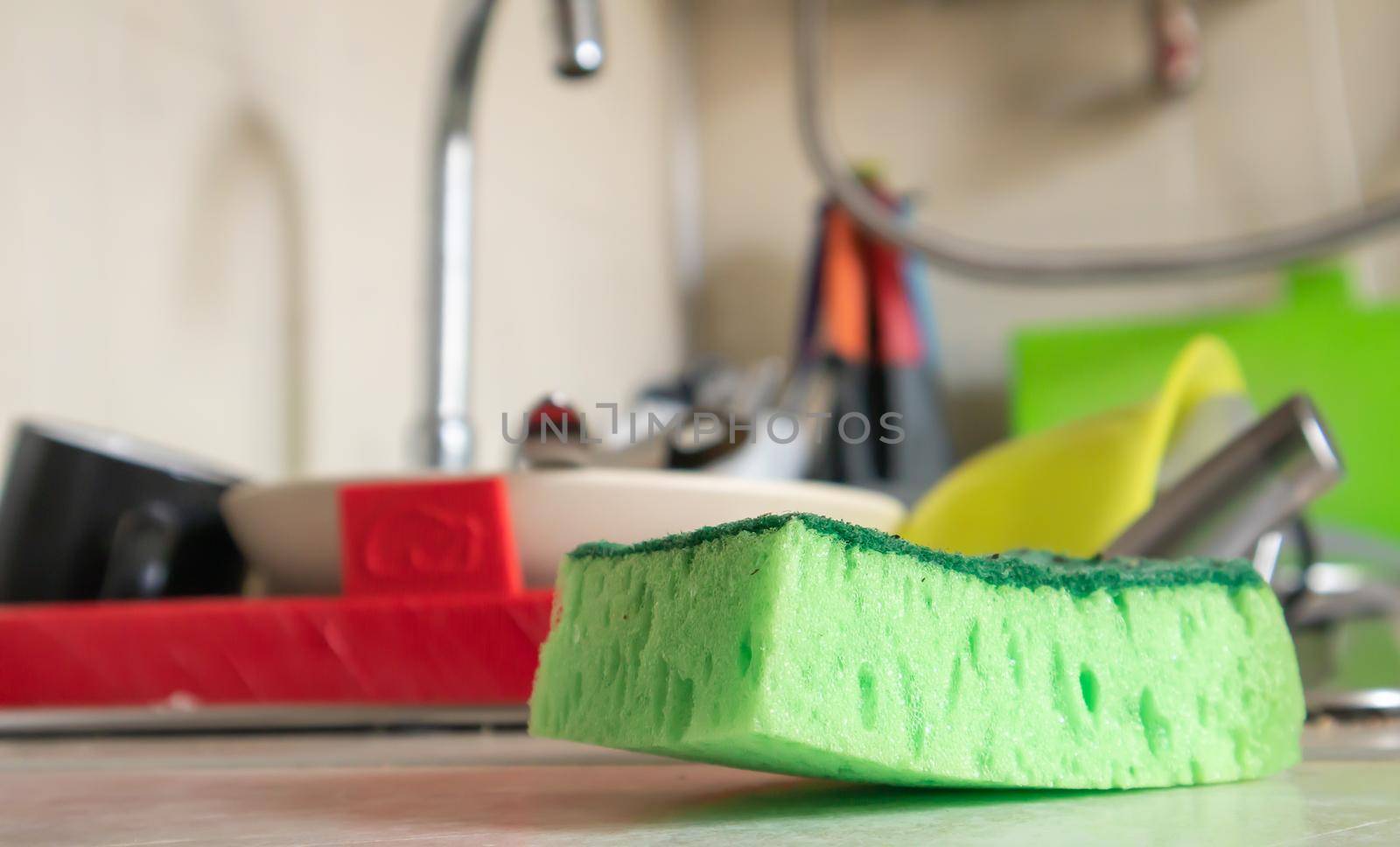 Green sponge and liquid soap dispenser for washing dishes on a dirty sink completely with dishes and kitchen utensils. Washing dishes in the kitchen by hand with detergent and a sponge