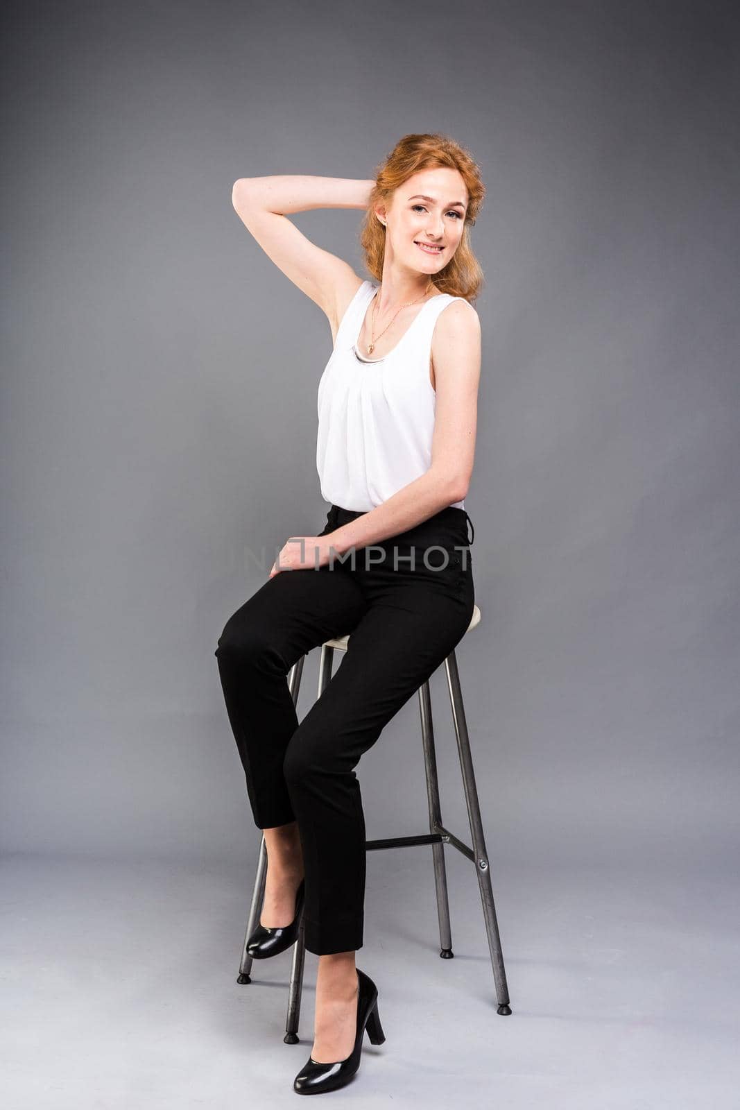 beautiful young caucasian woman with long red hair in high heels sitting on a chair, black trousers and white shirt in full growth on a gray background. Business concept by Tomashevska