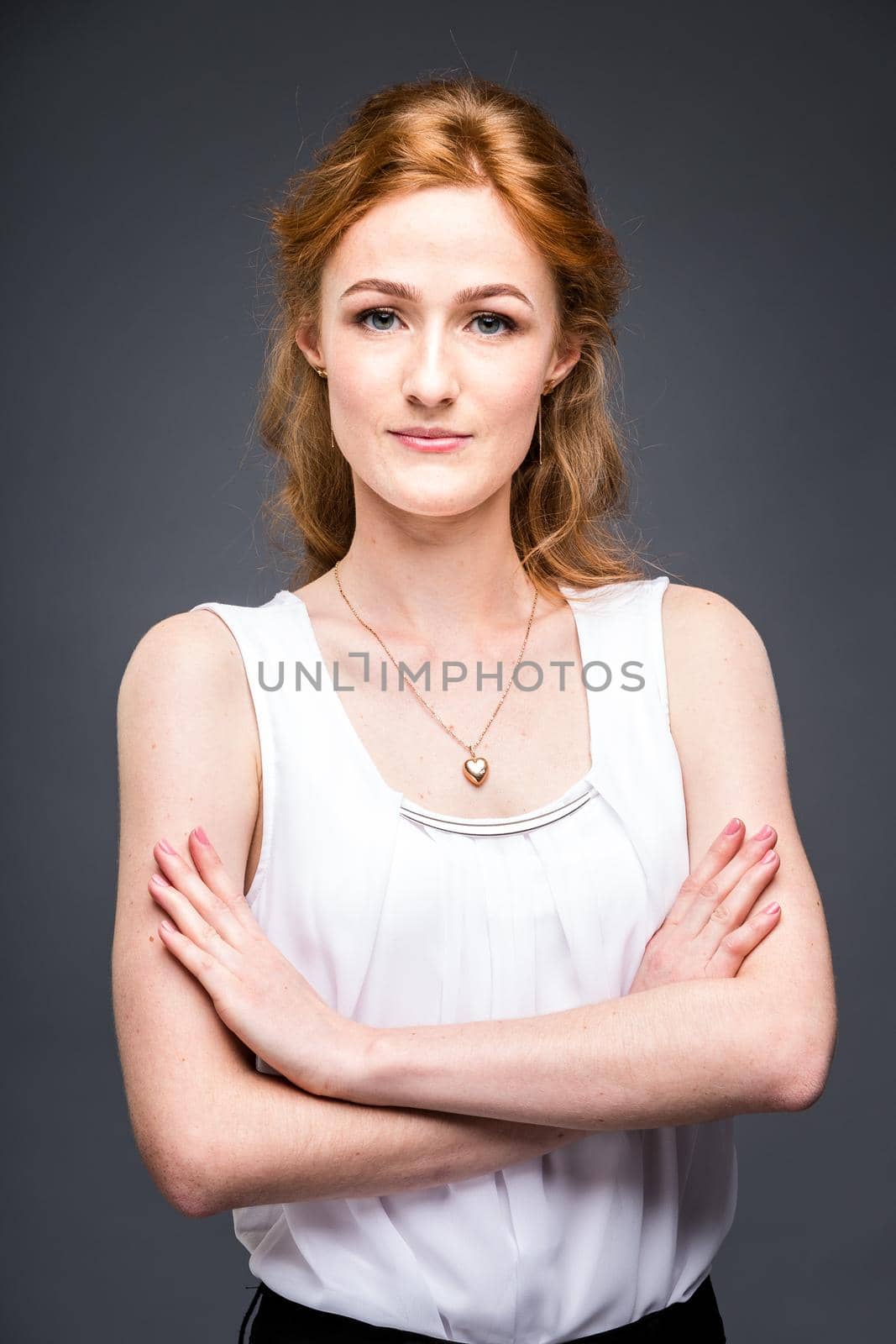 portrait of a young redhaired beautiful girl in the studio on a gray isolated background. A woman is standing with her arms folded and smiling in a white shirt with a short sleeve. Business concept by Tomashevska