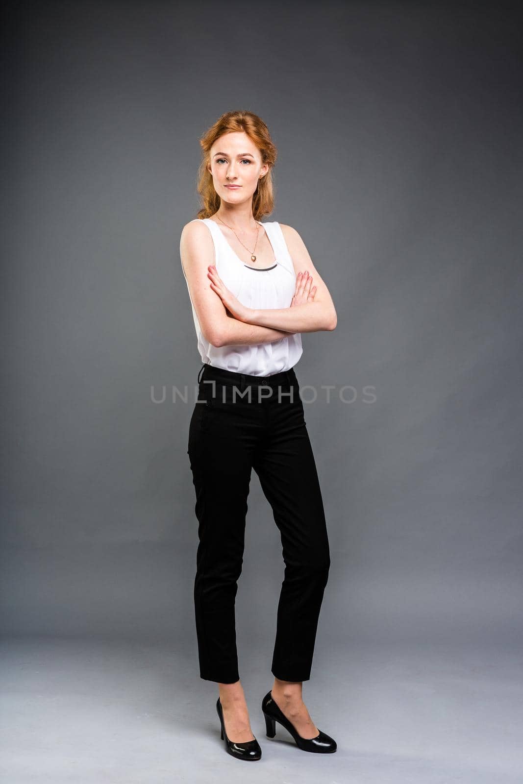 beautiful young Caucasian woman with long red hair in high heels, black trousers and a white shirt stands in full growth on a gray isolated background. by Tomashevska