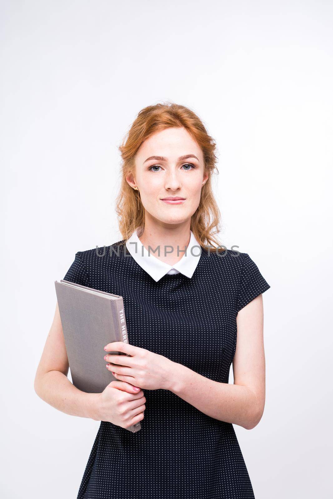 Beautiful girl with red hair and gray book in hands dressed in black dress on white isolated background.