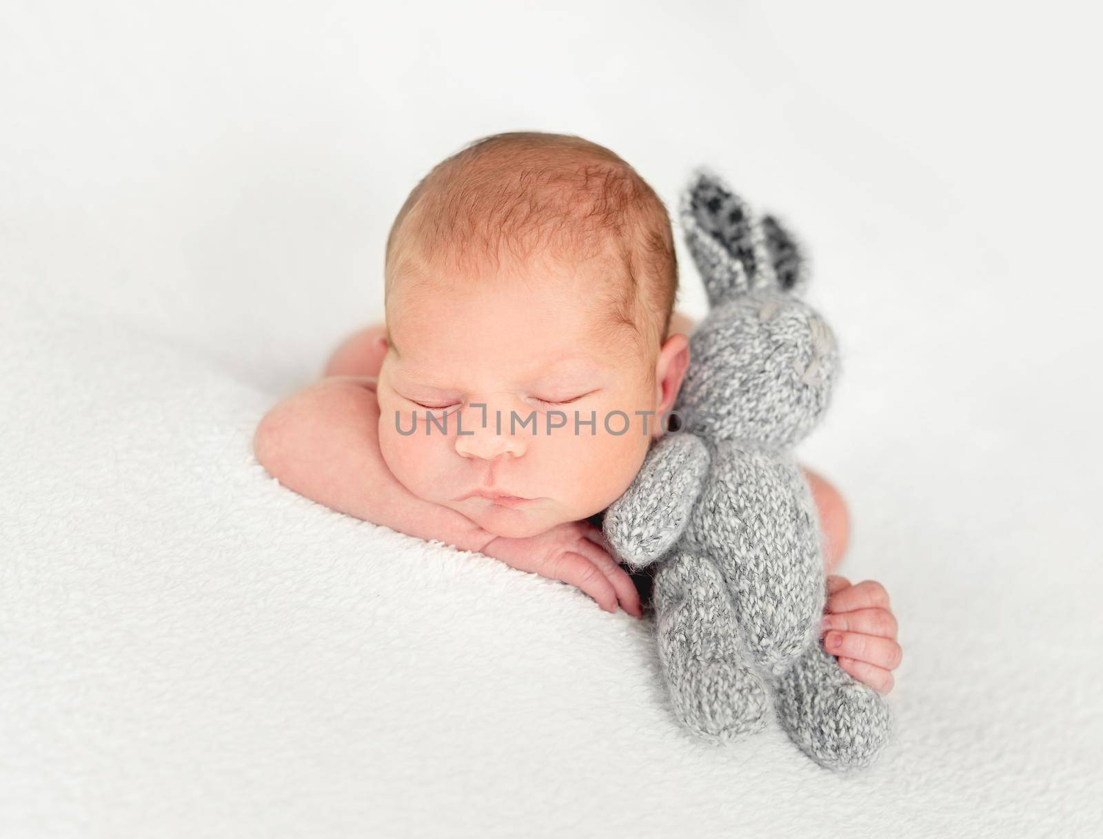 Baby embracing gray knitted hare toy, laying on white blanket background