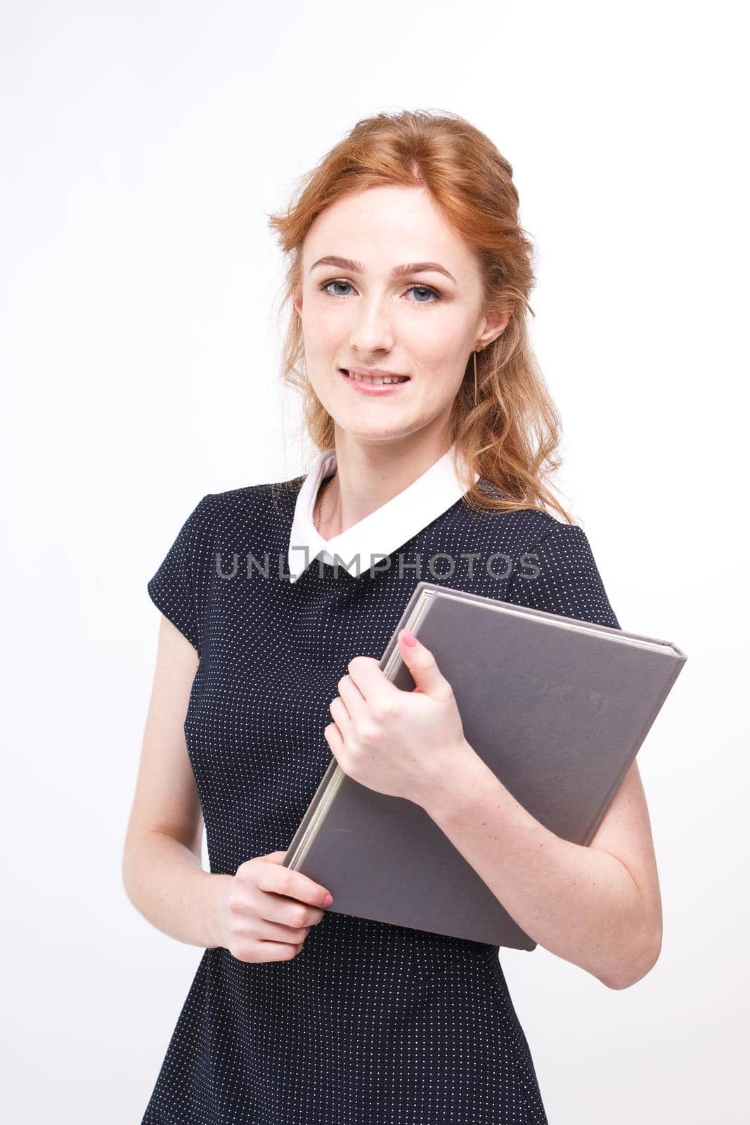 Beautiful girl with red hair and gray book in hands dressed in black dress on white isolated background.
