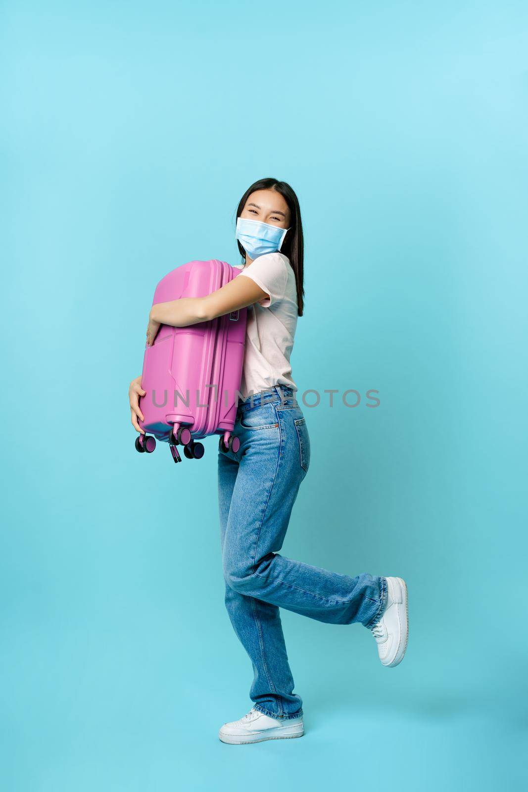 Full length of happy asian female tourist, dancing with suitcase in medical face mask, going on vacation, travelling with briefcase, standing over blue background.