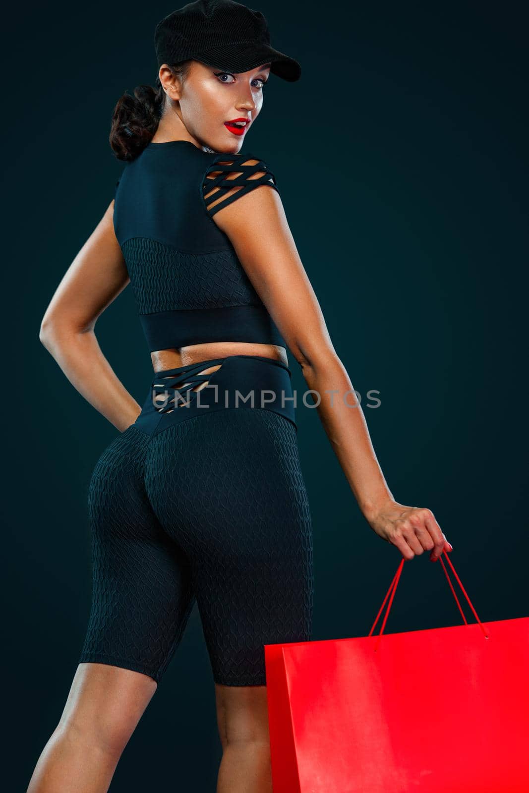 Black friday sale concept for fitness shop. Shopping woman in sportswear and hat holding red bag isolated on dark background.