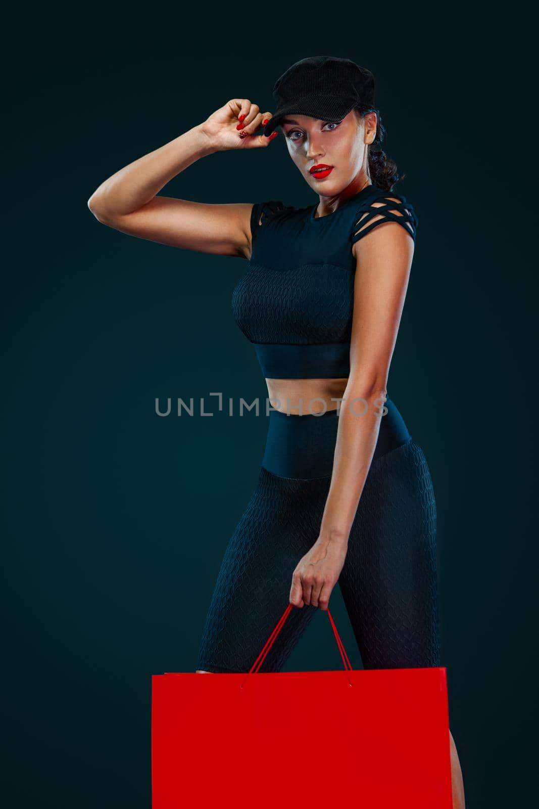 Black friday sale concept for fitness shop. Shopping woman in sportswear and hat holding red bag isolated on dark background by MikeOrlov