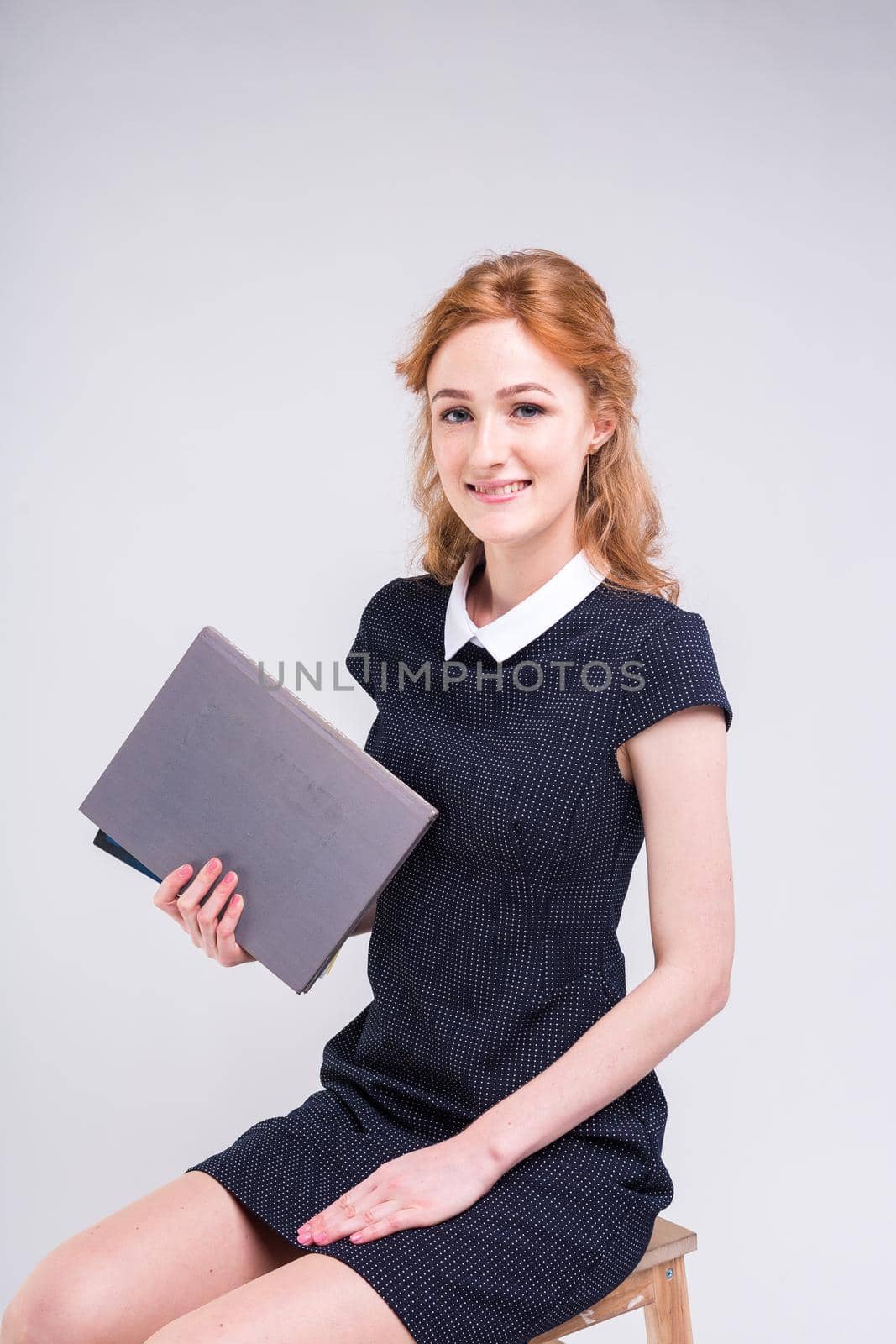 A young beautiful Caucasian female student sits on a chair in the studio on a white background. She is wearing a dress and holding a lot of books in her hands. Theme training and knowledge by Tomashevska