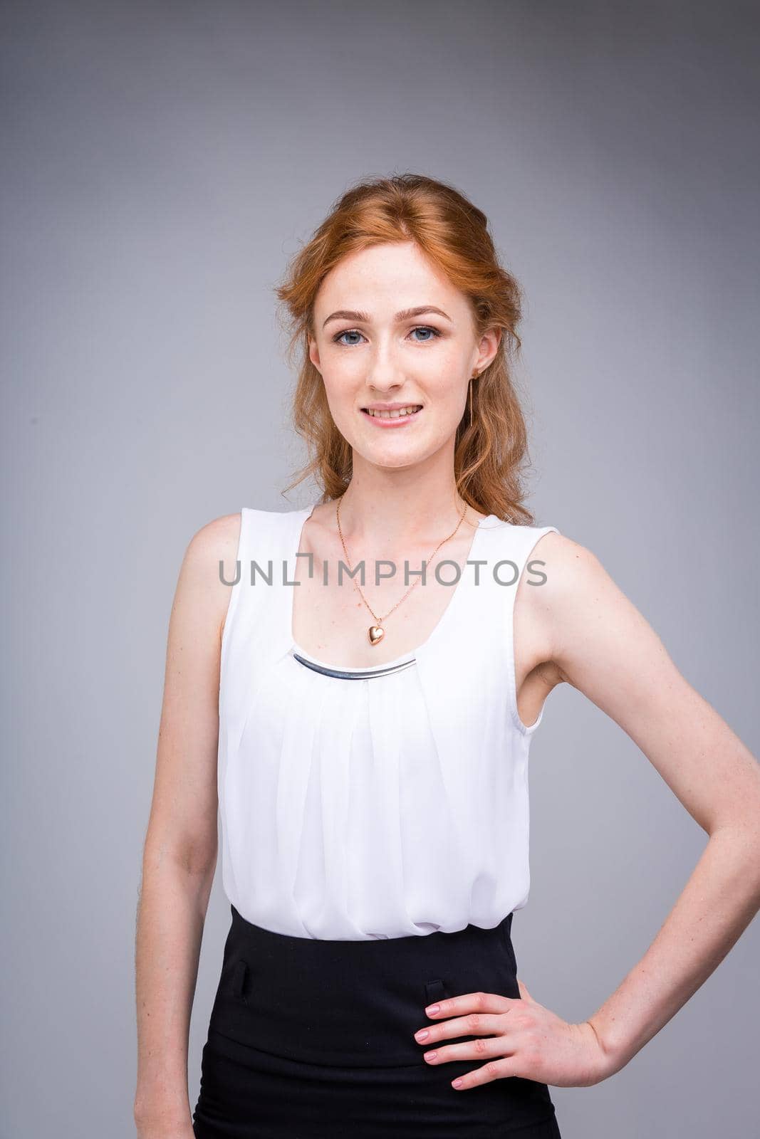 portrait vertical up to the waist young, beautiful business woman, student with lred, curly hair and freckles on gray background. SHe dressed in a white blouse with short sleeves about open shoulders.