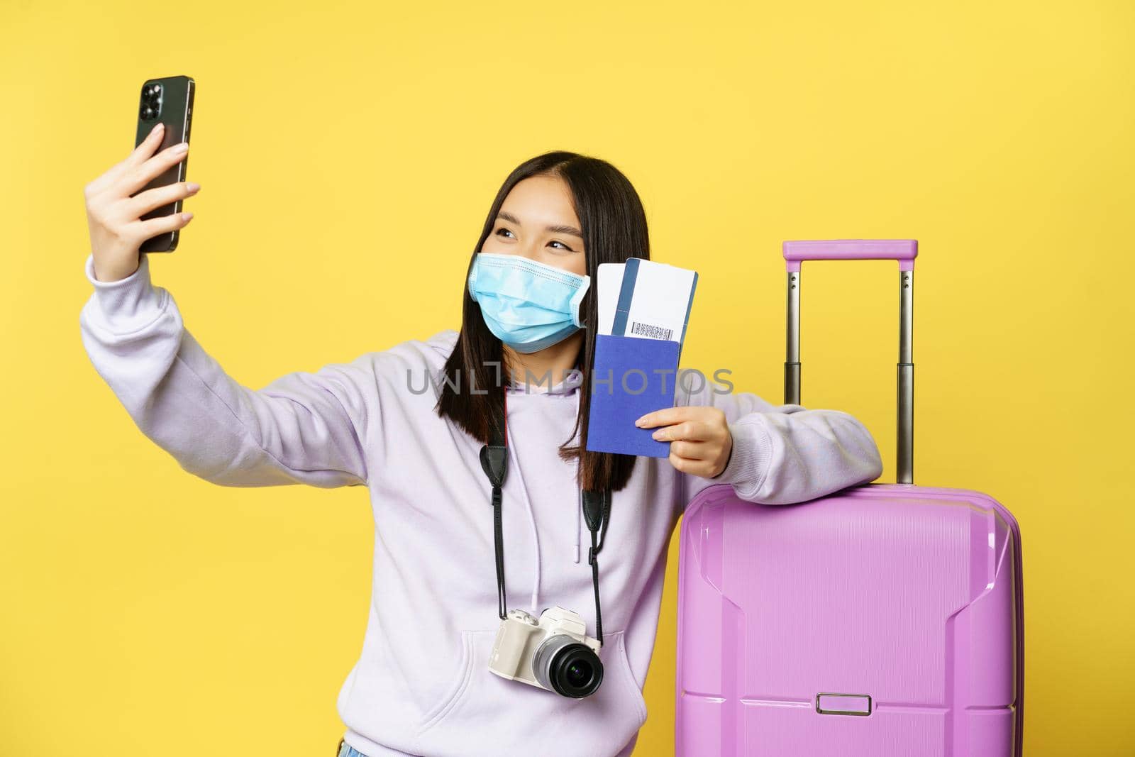 Happy asian woman taking selfie with passport and tickets near suitcase, photographing on smartphone in medical face mask, yellow background.