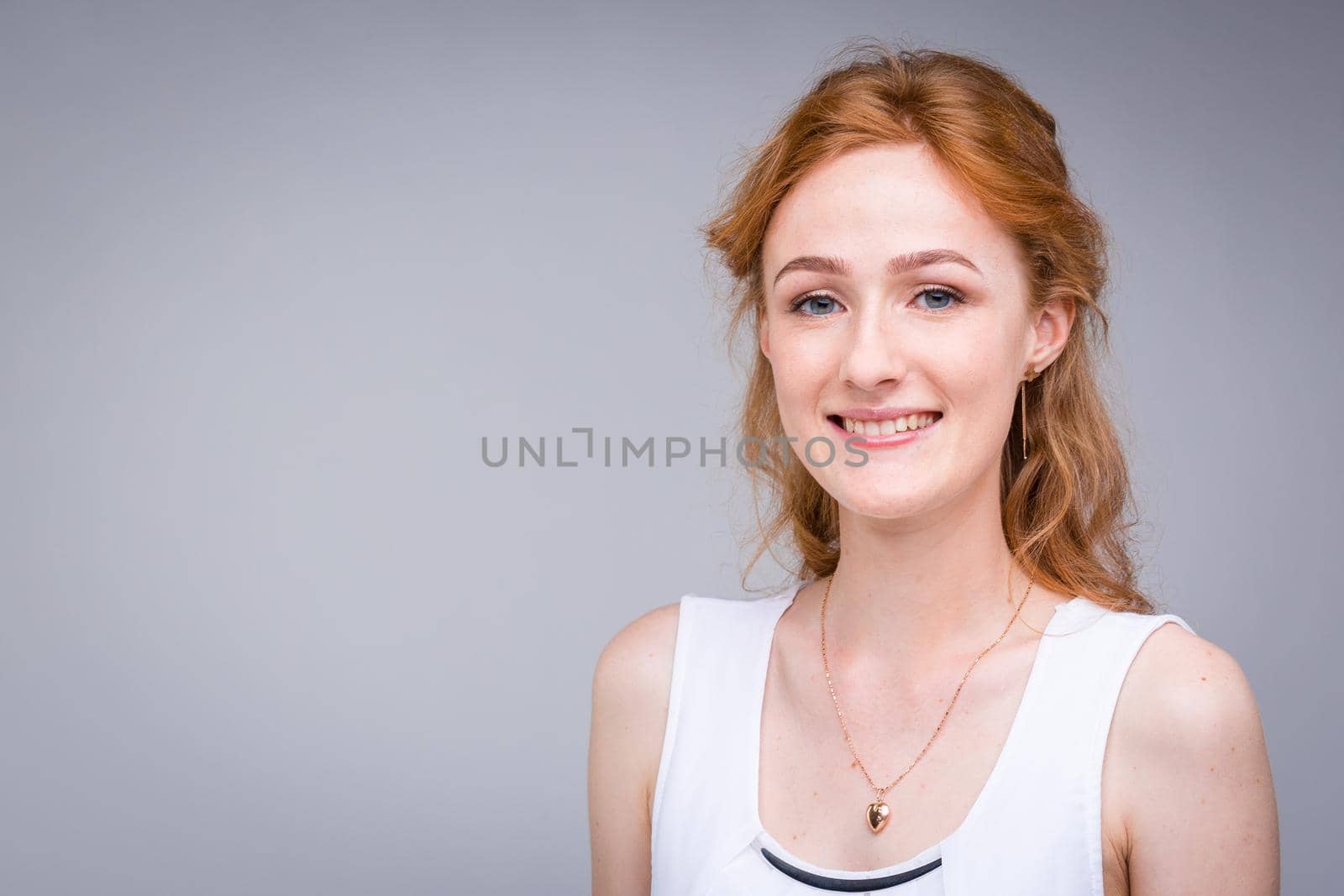 Closeup portrait young, beautiful business woman, student with lred, curly hair and freckles on face on gray background in the studio. Dressed in white blouse with short sleeves about open shoulders.