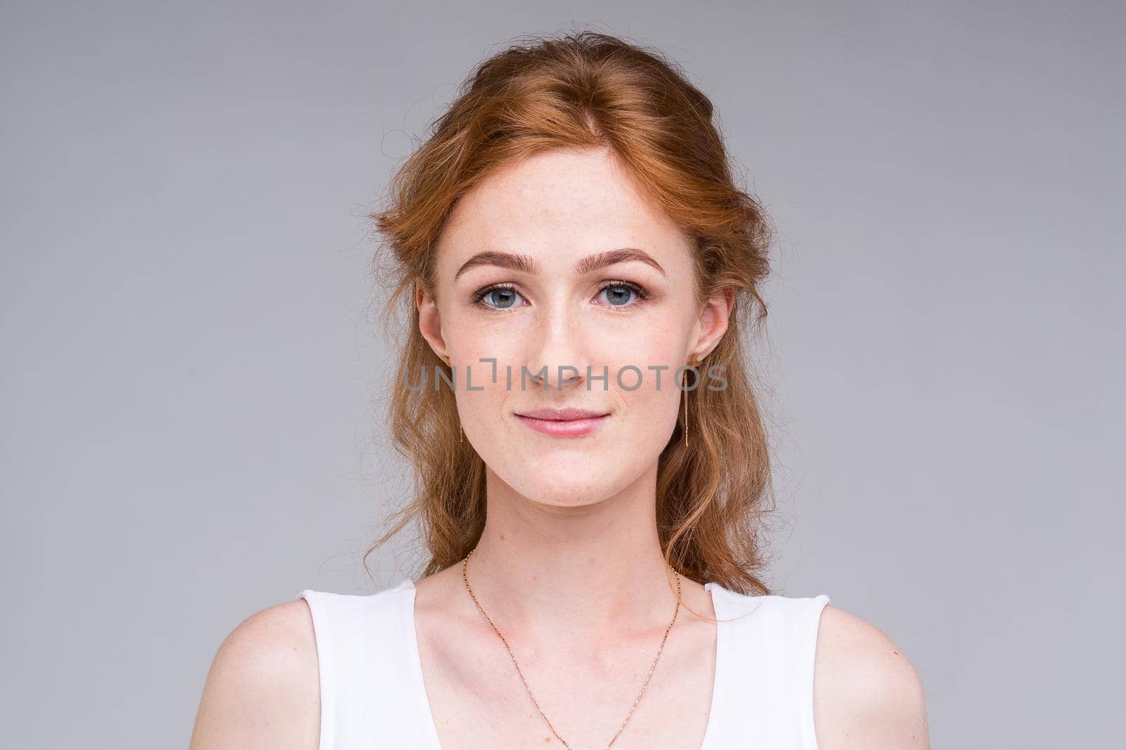 Closeup portrait young, beautiful business woman, student with lred, curly hair and freckles on face on gray background in the studio. Dressed in white blouse with short sleeves about open shoulders.