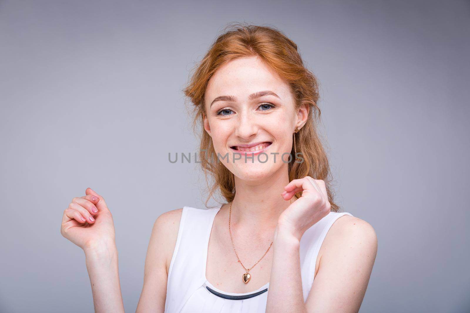 Closeup portrait young, beautiful business woman, student with lred, curly hair and freckles on face on gray background in the studio. Dressed in white blouse with short sleeves about open shoulders.