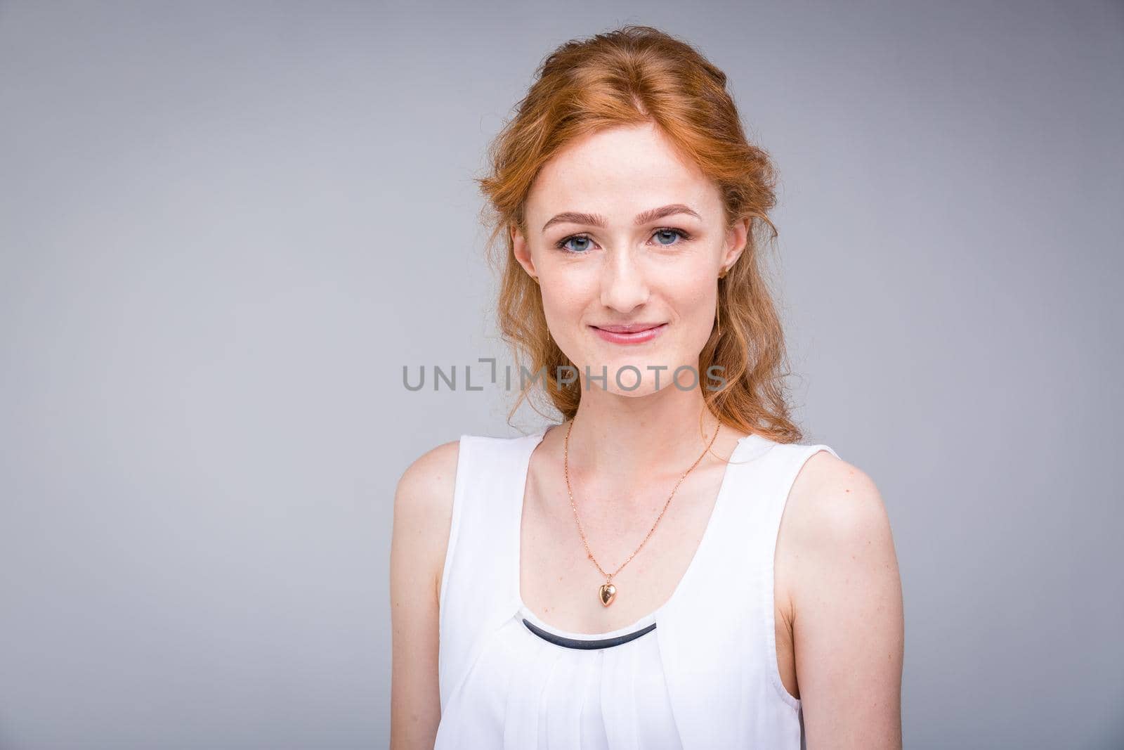 Closeup portrait young, beautiful business woman, student with lred, curly hair and freckles on face on gray background in the studio. Dressed in white blouse with short sleeves about open shoulders.