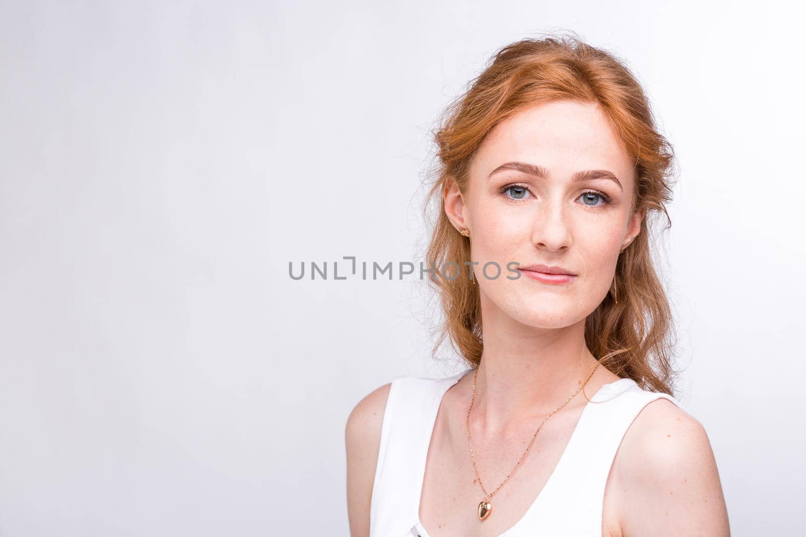 Portrait of a beautiful young woman of European, Caucasian nationality with long red hair and freckles on her face posing on a white background in the studio. Close-up student girl in a white blouse by Tomashevska