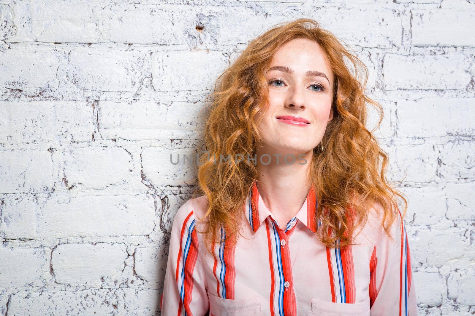 portrait of a beautiful beautiful young woman student with red curly hair and freckles on her face is leaning against a brick wall of gray color. Dressed in a red striped shirt.