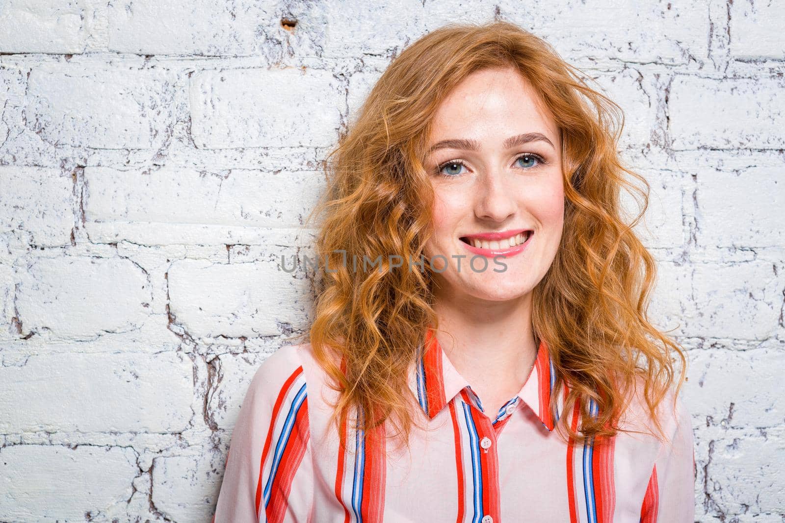 portrait of a beautiful beautiful young woman student with red curly hair and freckles on her face is leaning against a brick wall of gray color. Dressed in a red striped shirt.