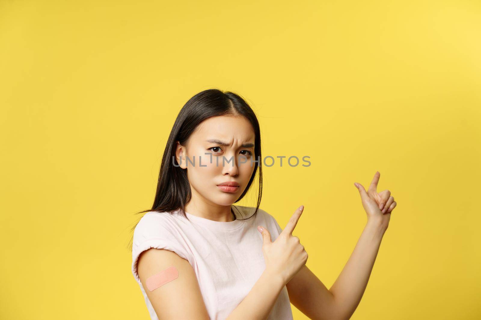 Covid-19 vaccination concept. Disappointed sulking asian girl pointing at promo banner, being vaccinated from coronavirus, yellow background.