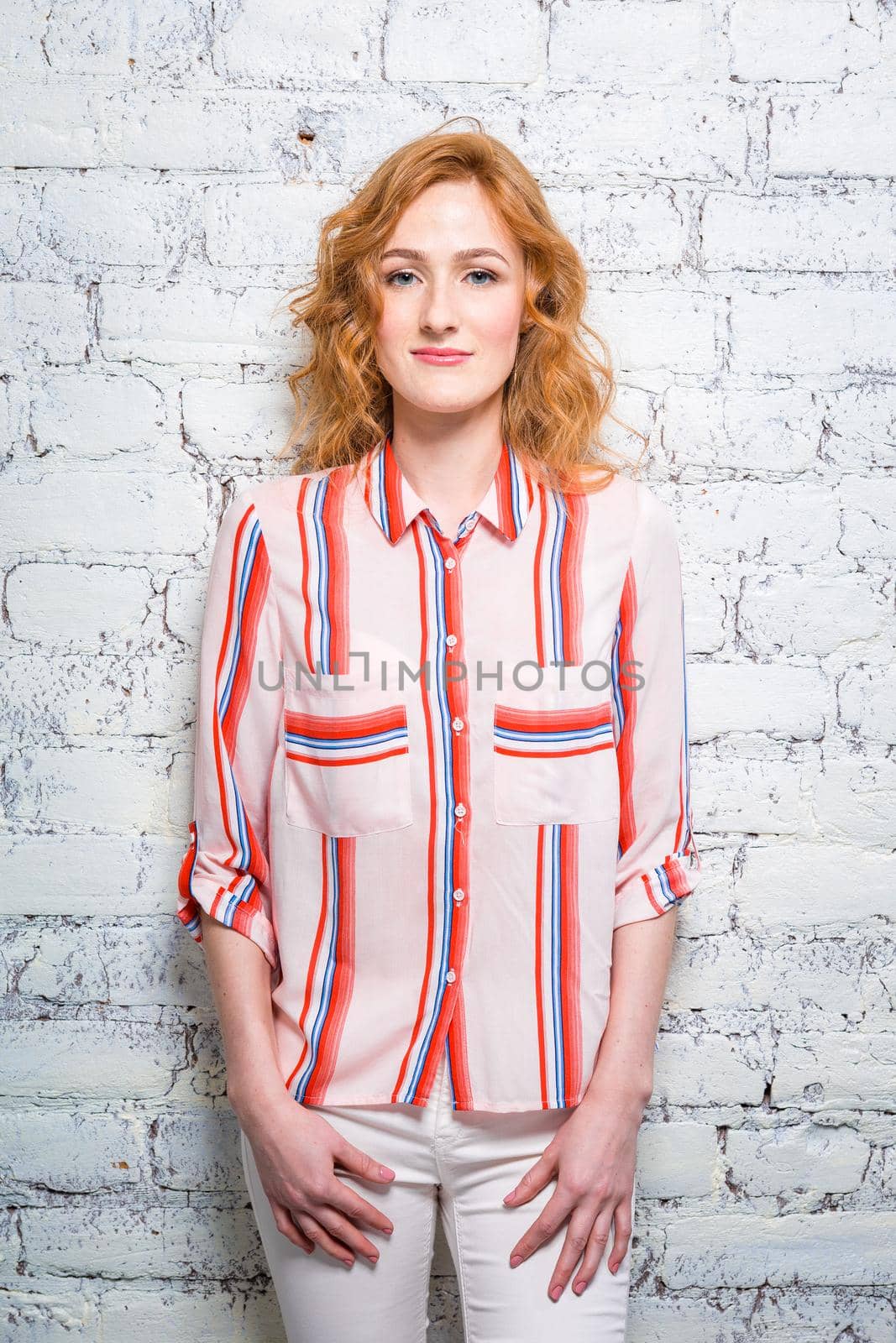 A beautiful young woman student with red curly hair and freckles is leaning against a brick wall of gray color. Dressed in a red striped shirt and white pants.