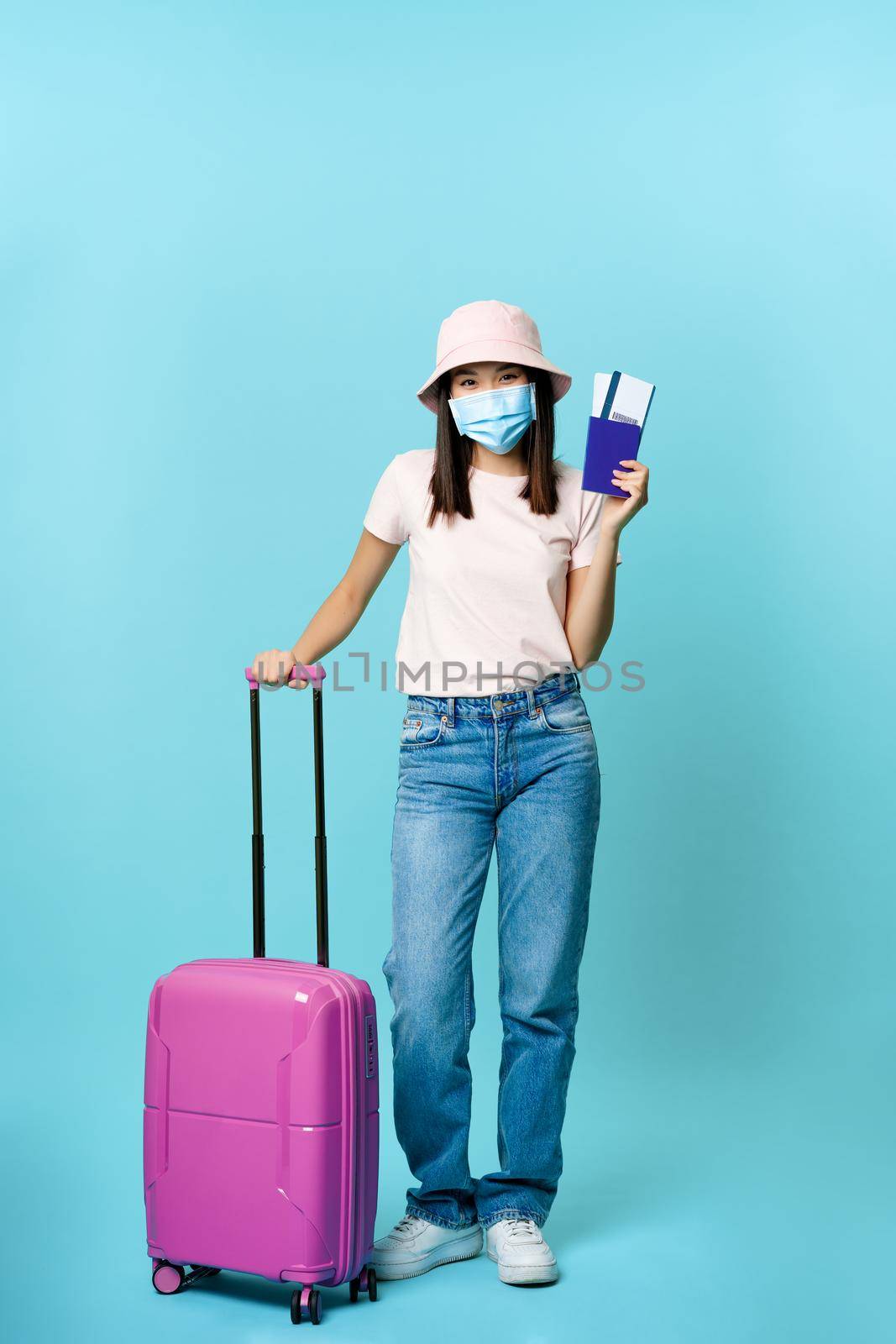 Vertical shot of happy asian woman with suitcase, showing passport and two tickets, wearing face mask from coronavirus, blue background.