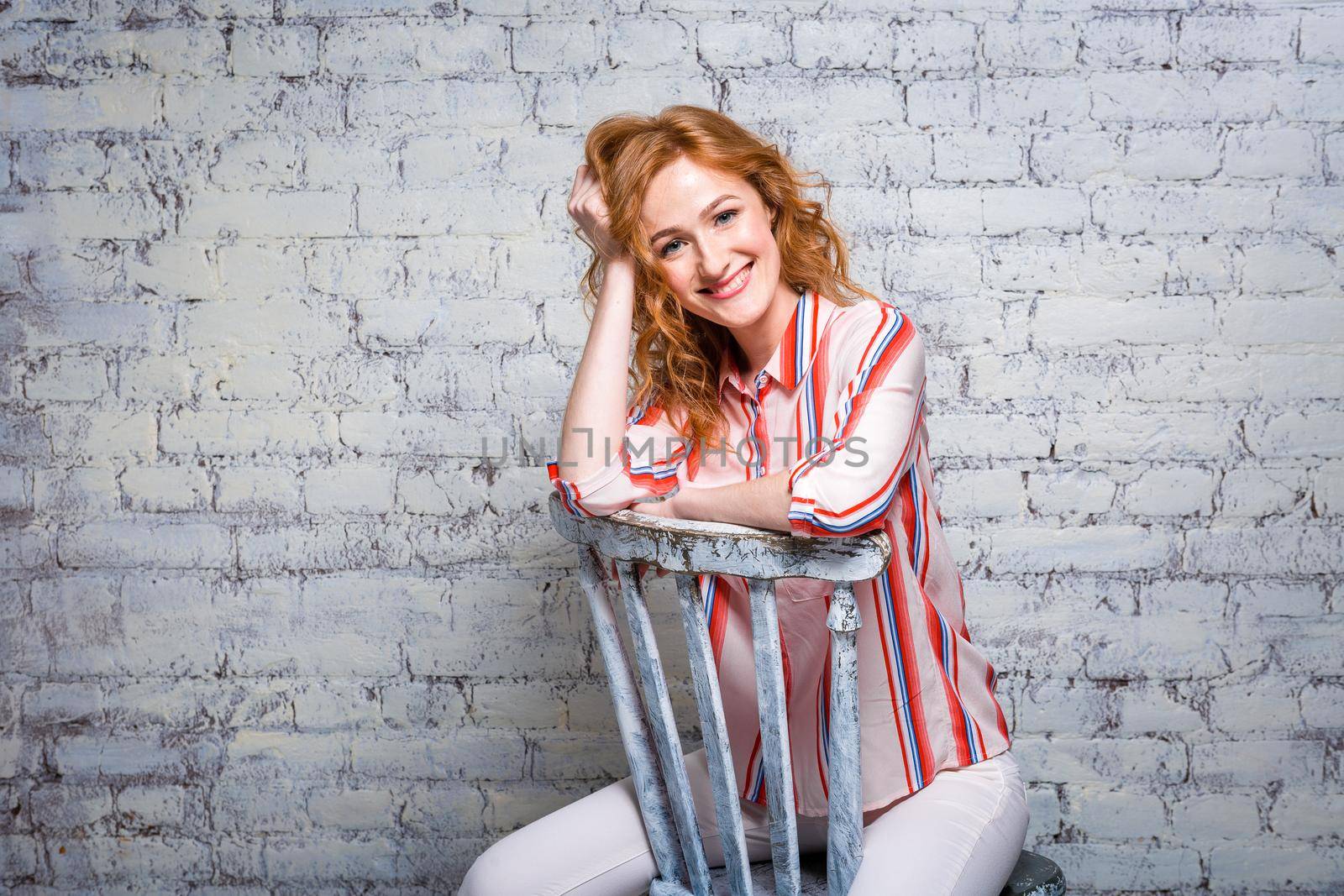 close-up portrait Beautiful young female student with red curly hair and freckles on her face sitting on a brick wall in gray background. Dressed in a red striped shirt. Hands touching the hair.
