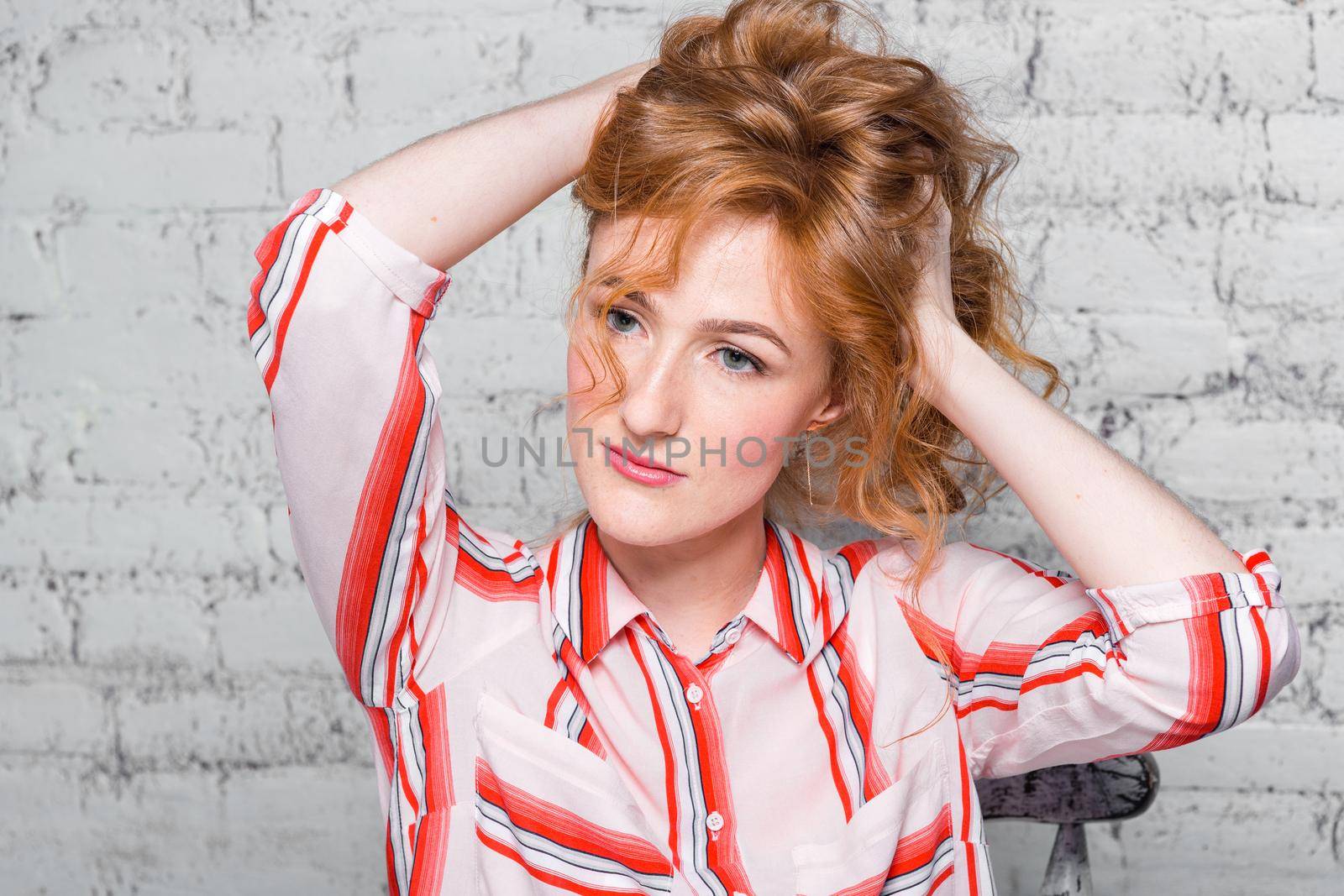 close-up portrait Beautiful young female student with red curly hair and freckles on her face sitting on a brick wall in gray background. Dressed in a red striped shirt. Hands touching the hair by Tomashevska