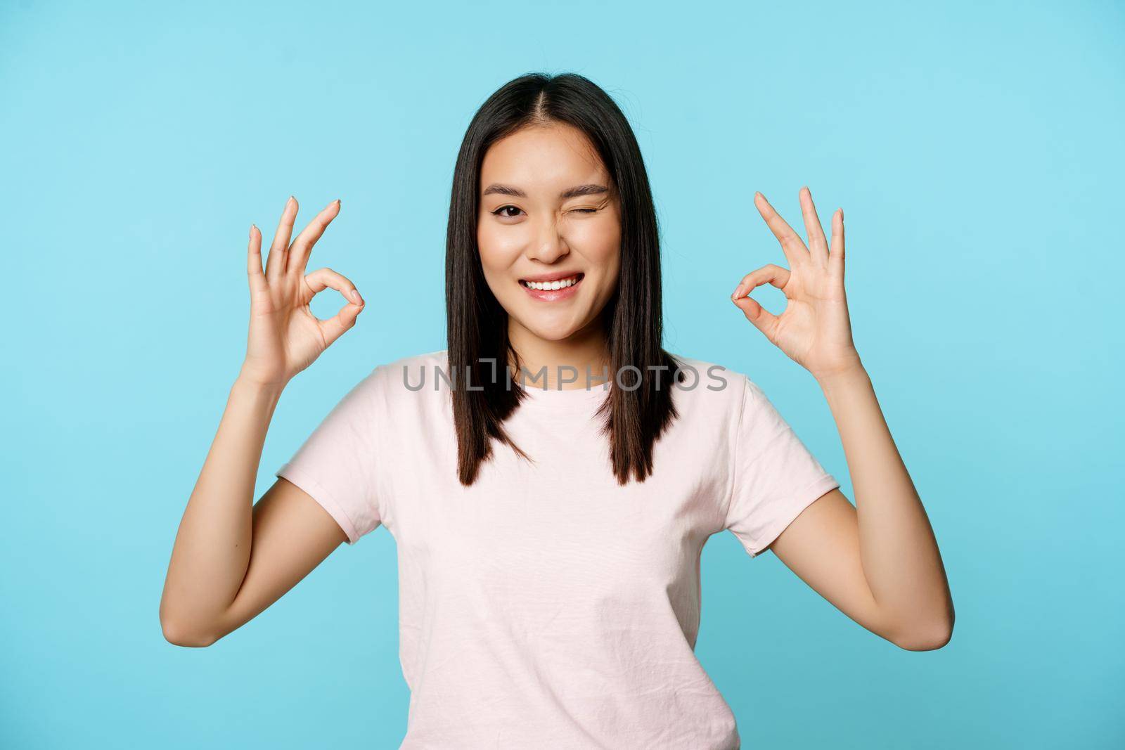 Smiling korean woman winking, showing okay signs, recommending company or store, standing in t-shirt over blue background by Benzoix