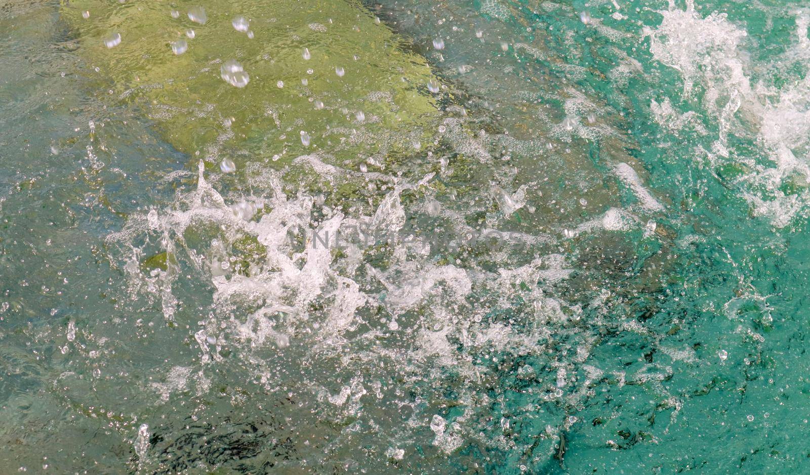 Frequent drops of water in a fountain in slow motion fall on a clean water surface with splashes and bubbles. Jets of water sparkle in the sun. The spray slowly spreads to the sides. Close-up