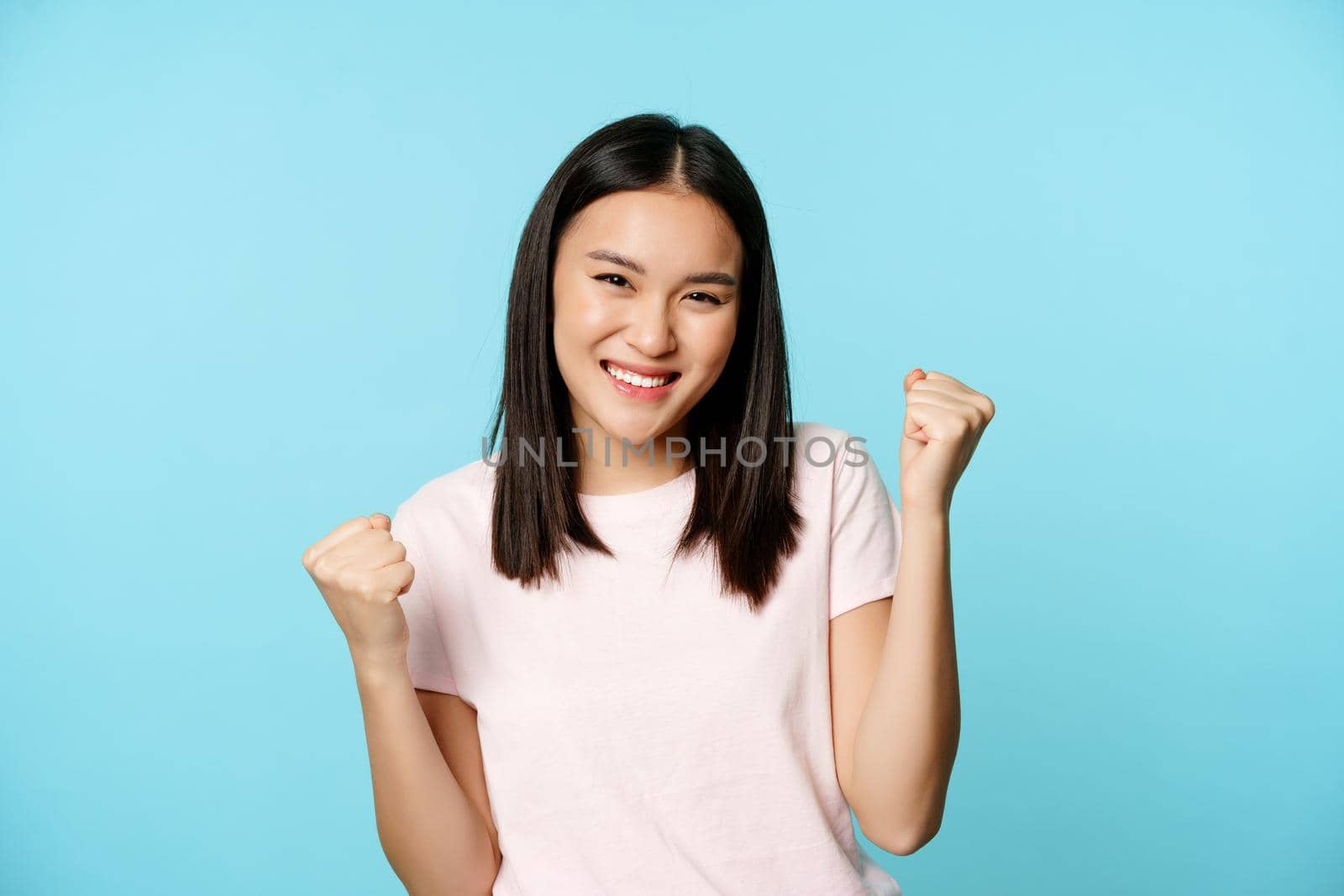 Enthusiastic cute asian girl winning, rejoicing from great news, victory dance, smiling pleased, standing over blue background.