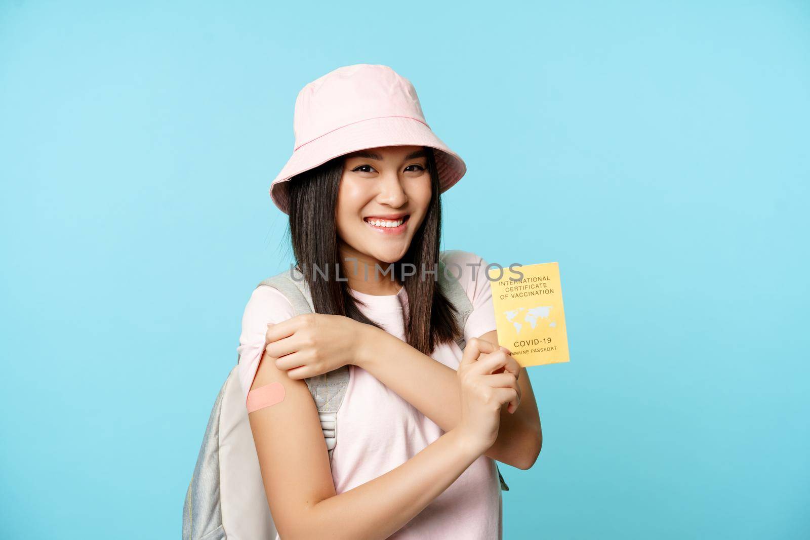 Smiling korean tourist, happy woman shows her arm vaccinated arm and covid-19 international vaccination certificate, standing over blue background by Benzoix