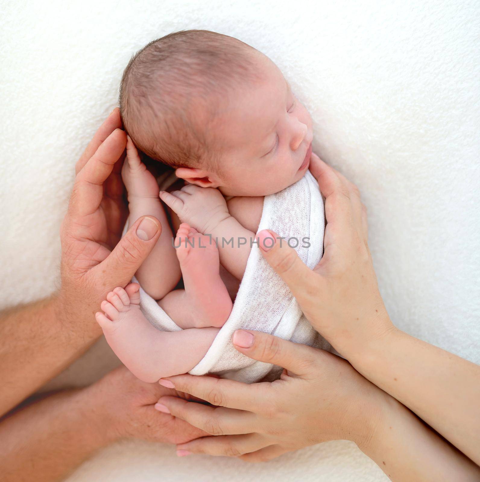 Parents holding newborn cute child, wrapped in white blanket on the bed