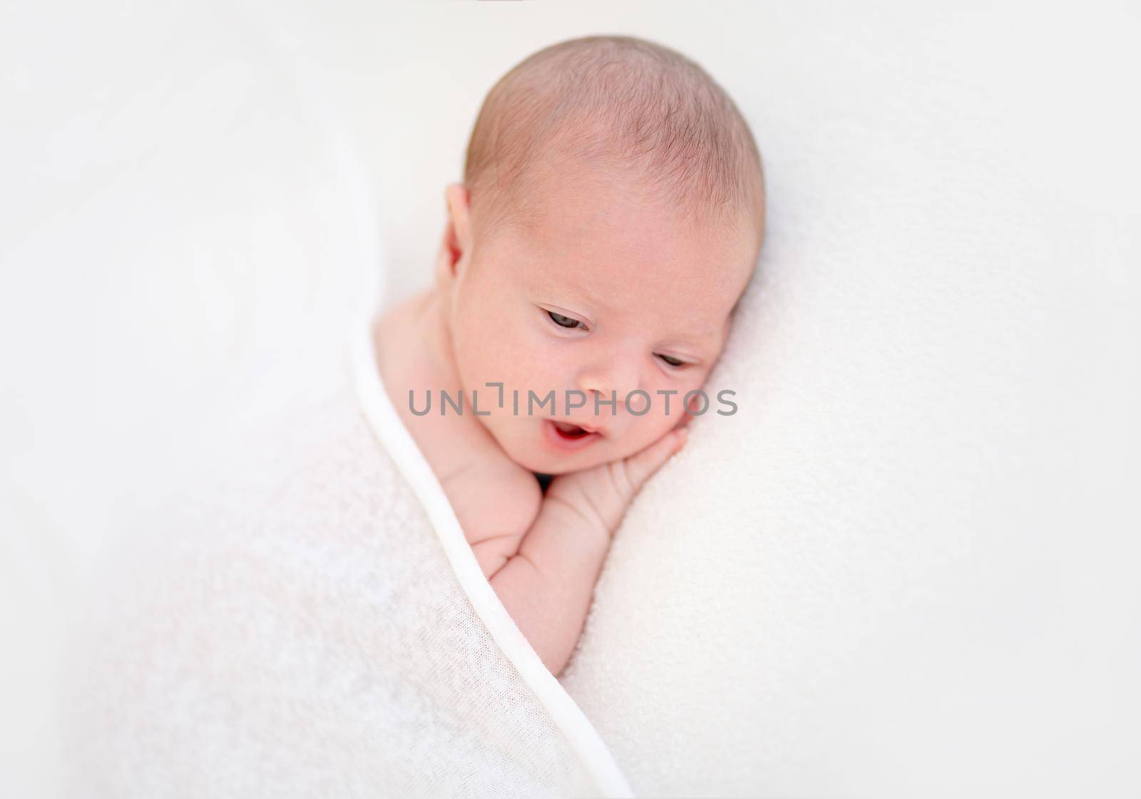 Closeup portrait of sleepy newborn kid, holding hand under head, lying on bed on white background