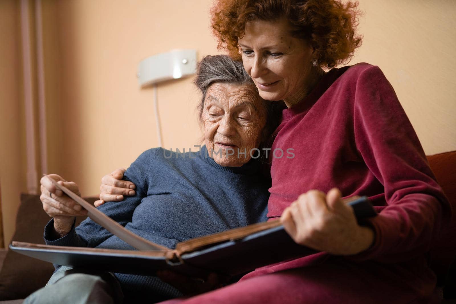 Mature daughter visits senior mother and has fun watching family photo album, sitting in embrace in living room couch. Mothers Day. Elderly woman and old gray hair grandmother laugh and remember youth