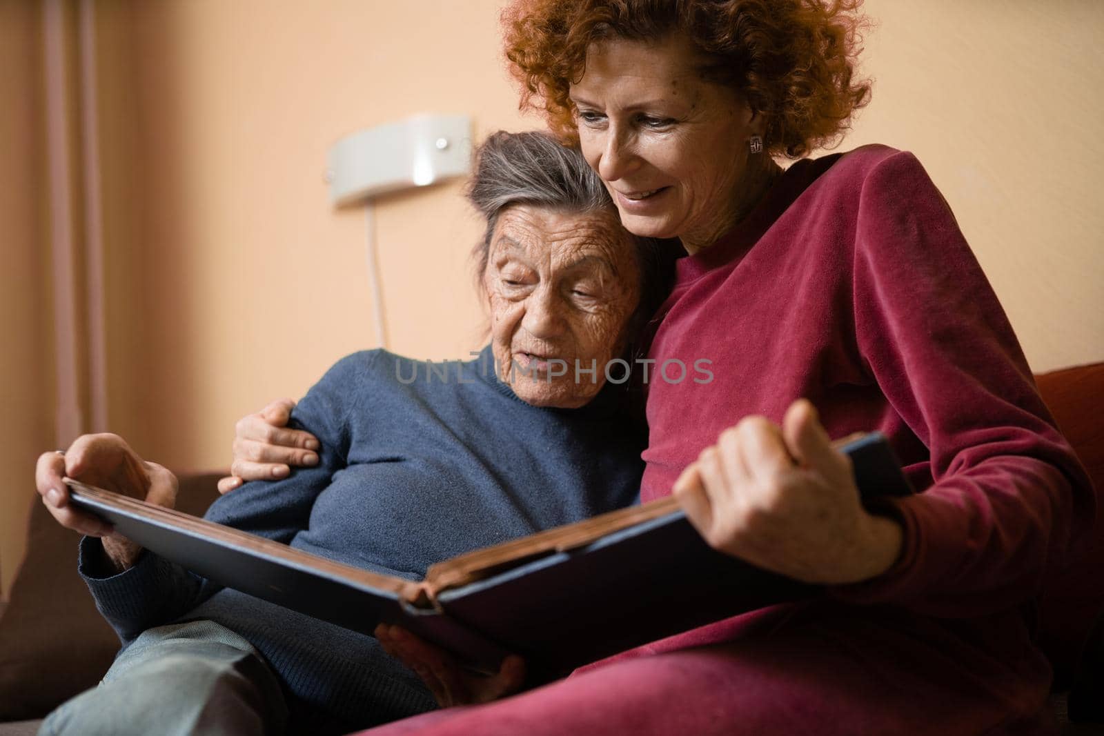 Senior woman and her adult daughter looking at photo album together on couch in living room, talking joyful discussing memories. Weekend with parents, family day, thanksgiving, mom's holiday by Tomashevska