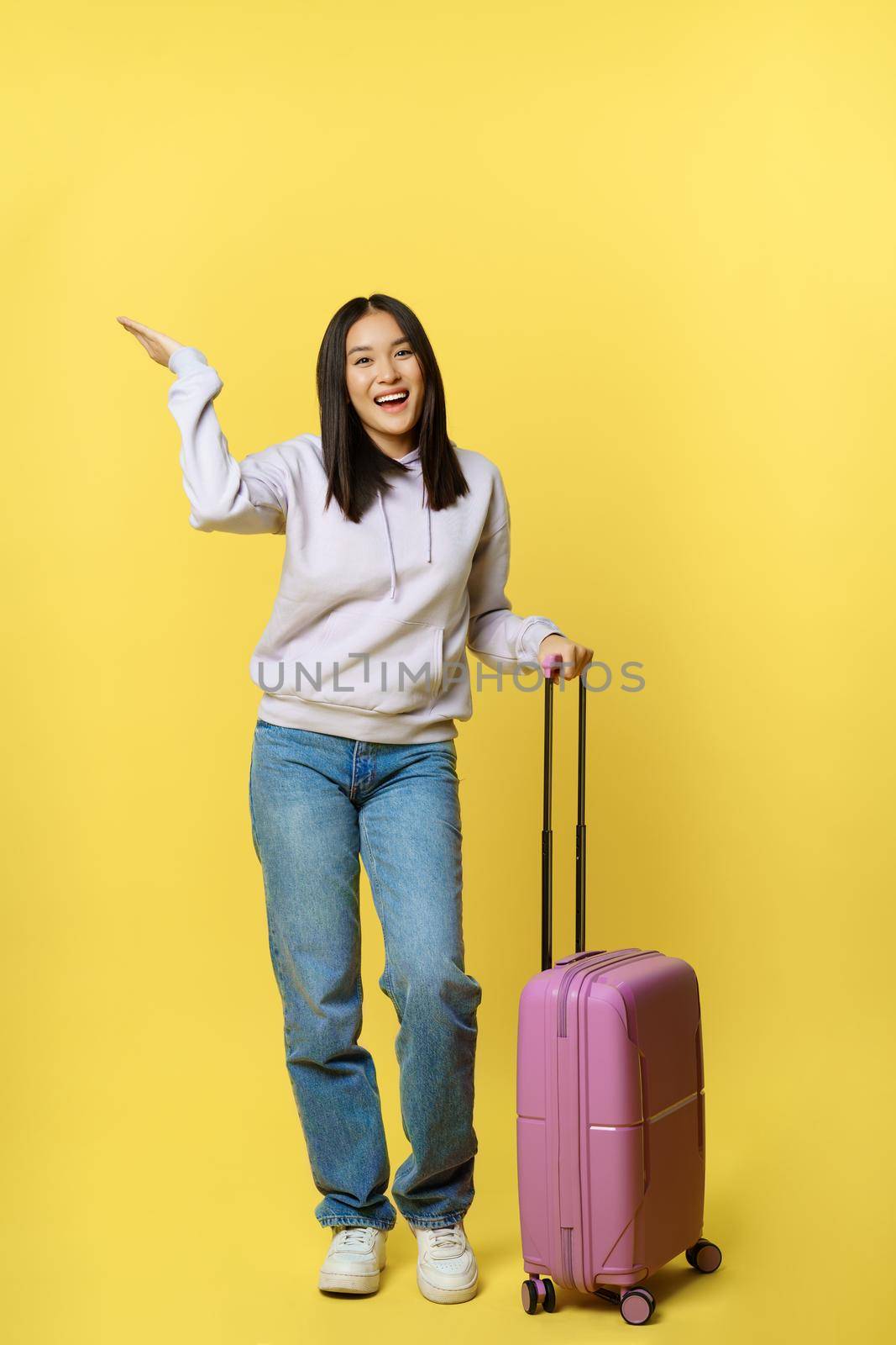 Full length shot happy asian girl going on vacation, tourist with suitcase smiling and looking upbeat, standing over yellow background.
