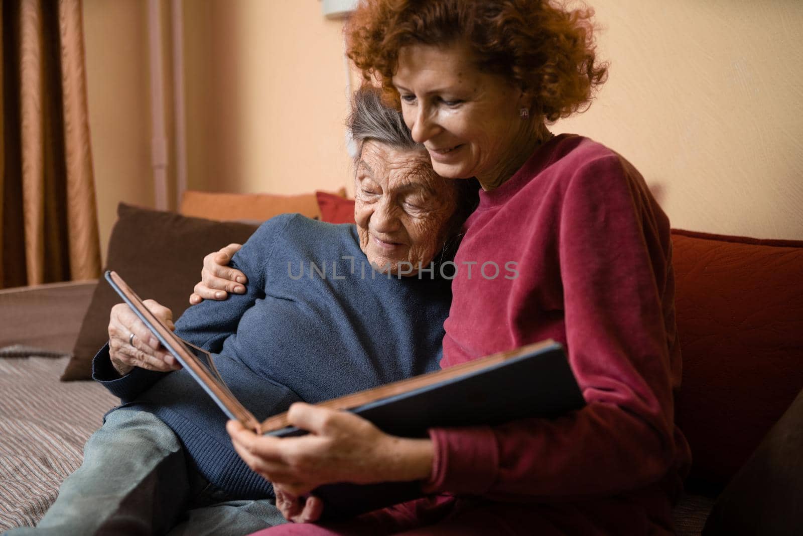 Positive aged ladies looking album photos sitting sofa at home, cheerful friends. Senior woman and her mature nurse watching photo album. Granny showing her daughter memories from the past.
