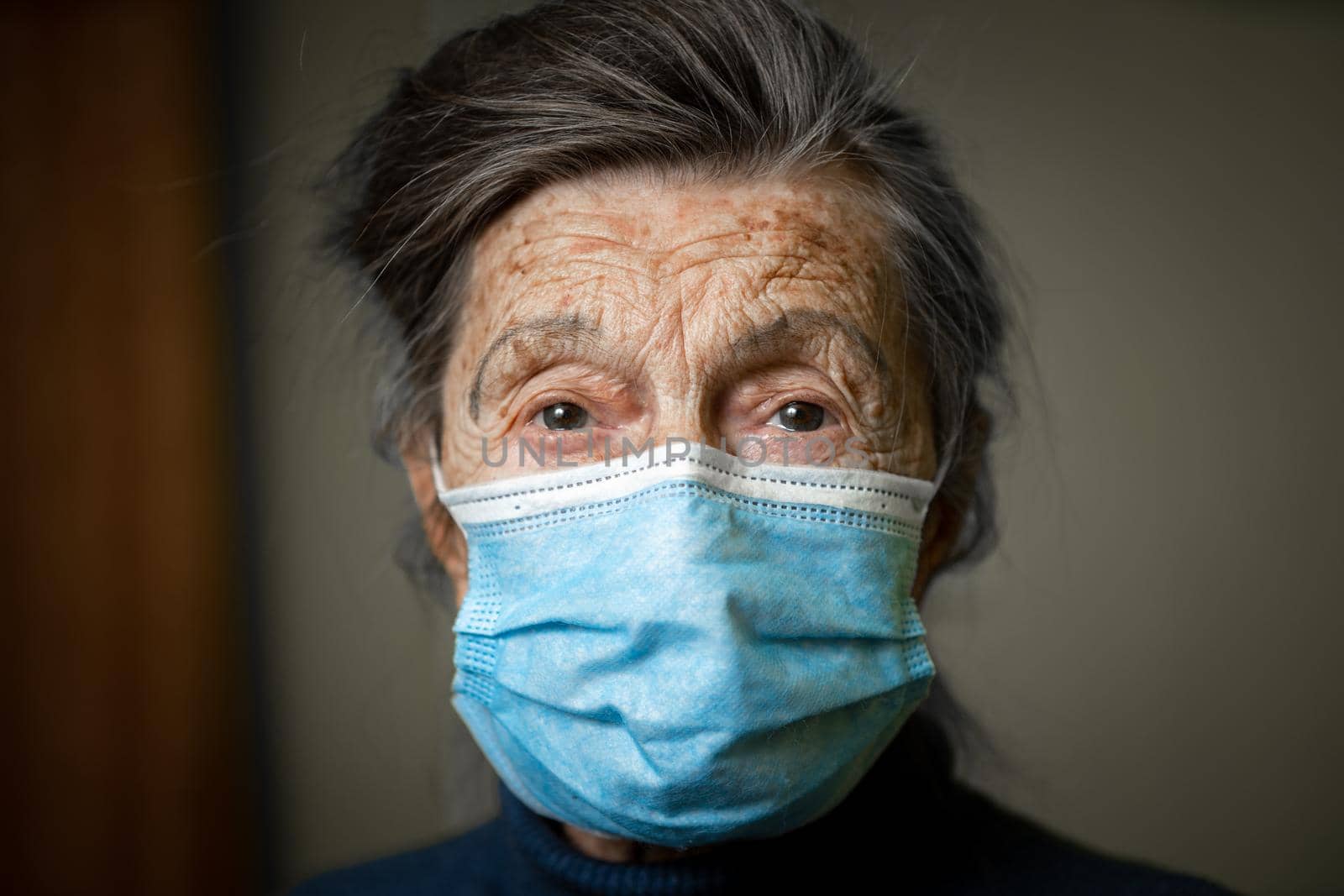Senior woman in medical mask posing looking in camera at home. Topic help and support for old people during coronavirus epidemic. Elderly female is lonely during lockdown covid 19 in quarantine by Tomashevska